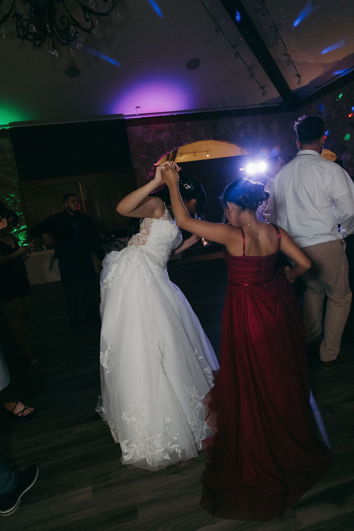 Bride in white gown dancing with bridesmaid in red dress under colorful lights at reception.