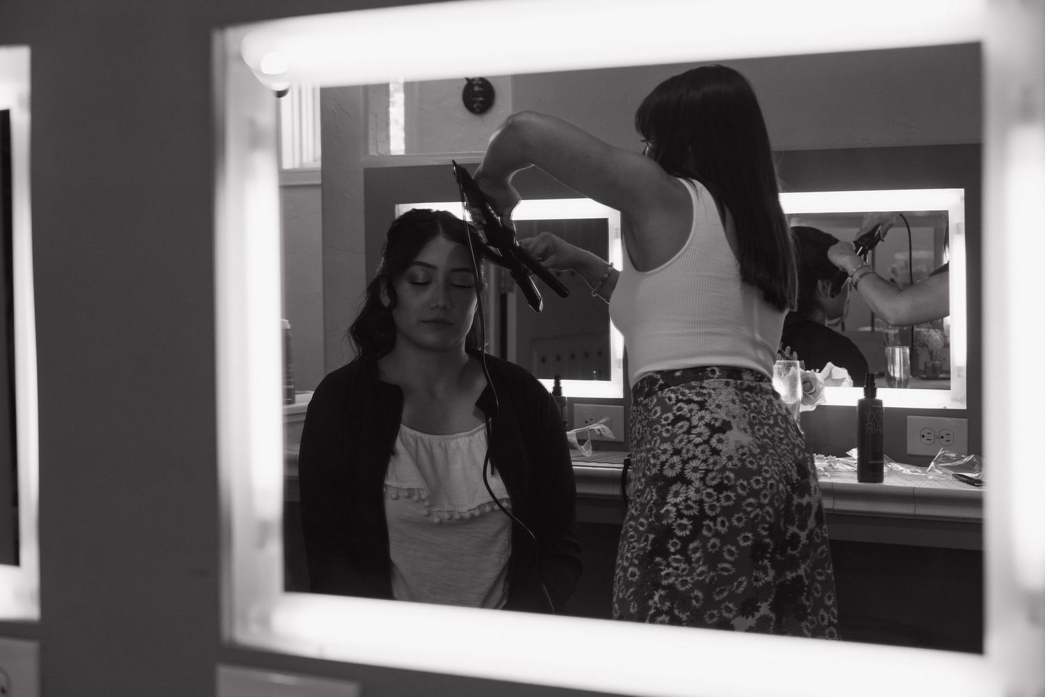 Black and white photo of bride getting hair styled in mirror reflection during wedding preparations.