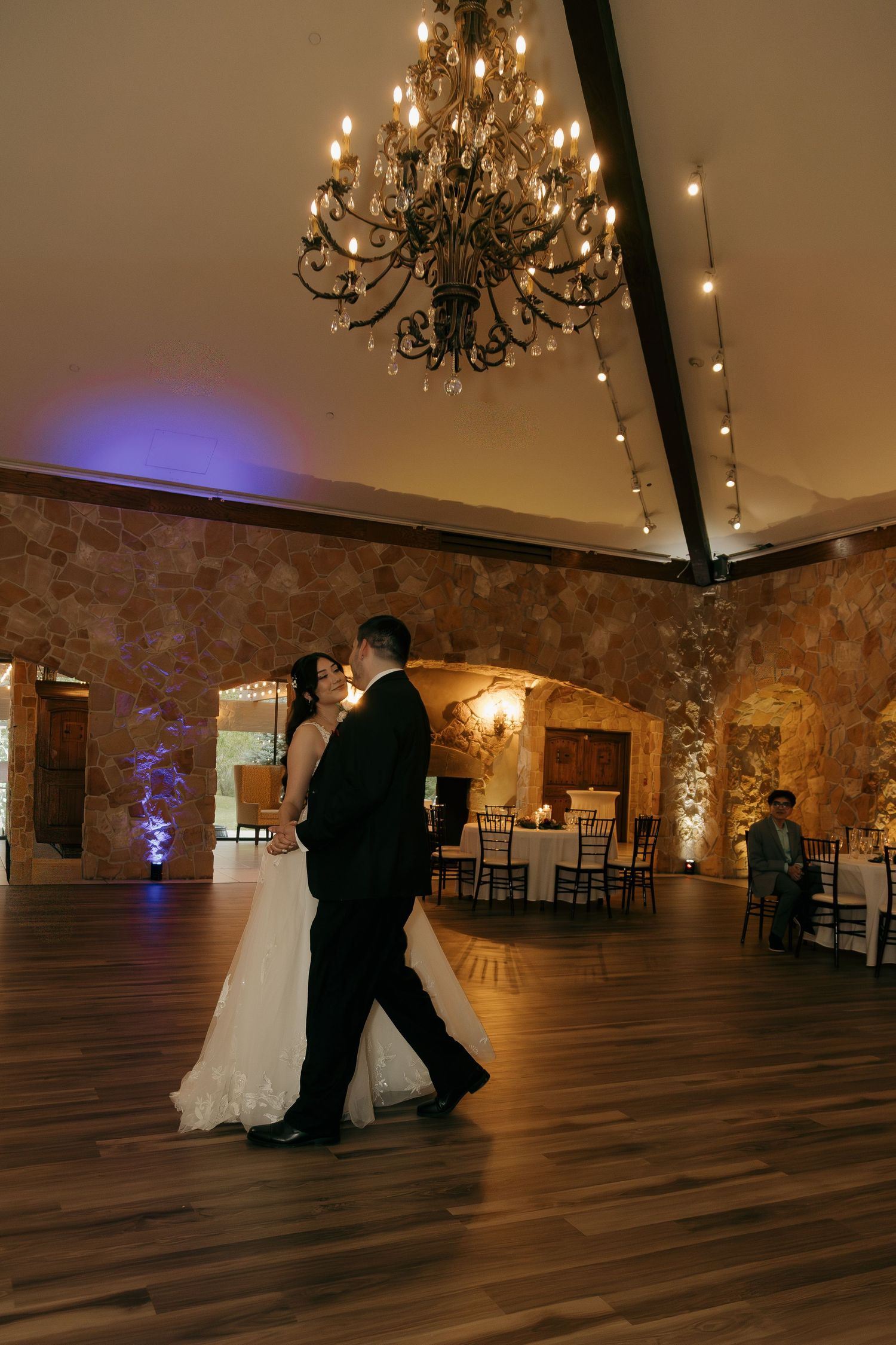 Bride and groom sharing their first dance under a grand chandelier in a rustic stone ballroom.