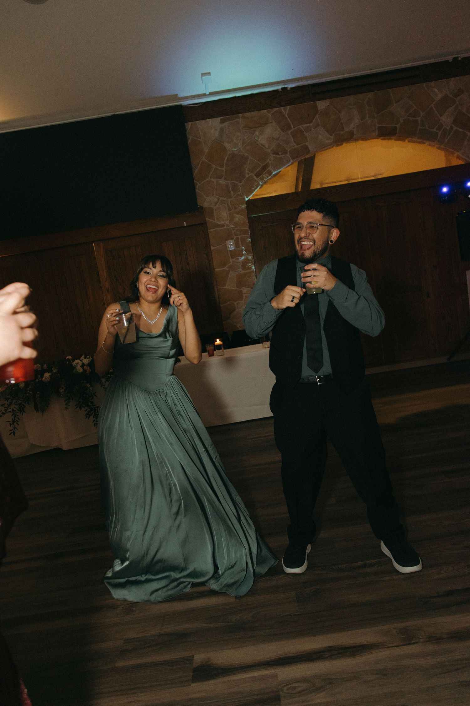 Man in vest and woman in sage green gown laugh and sing together on a wedding reception dance floor.