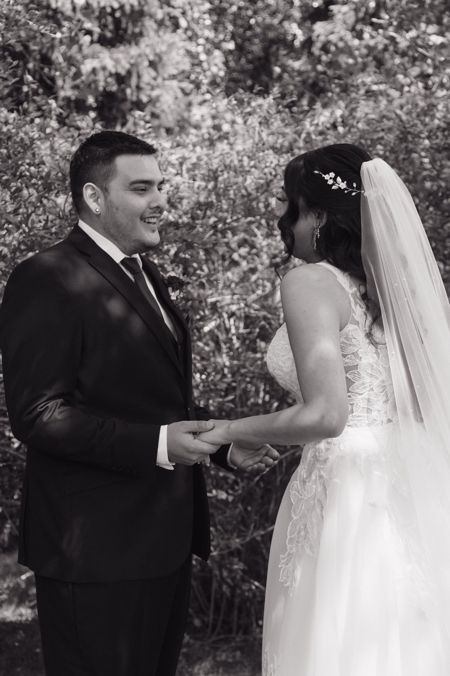 Black and white photo of smiling groom holding brides hands in wedding attire.