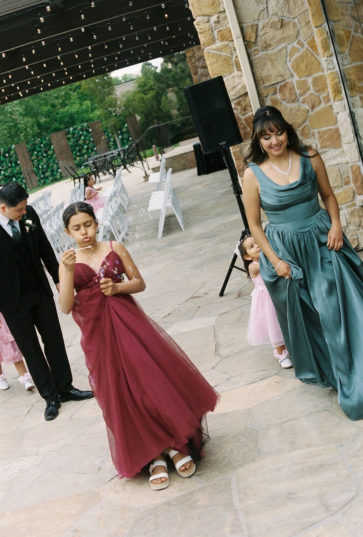 Young girl in burgundy gown and woman in teal dress walk at outdoor wedding reception.