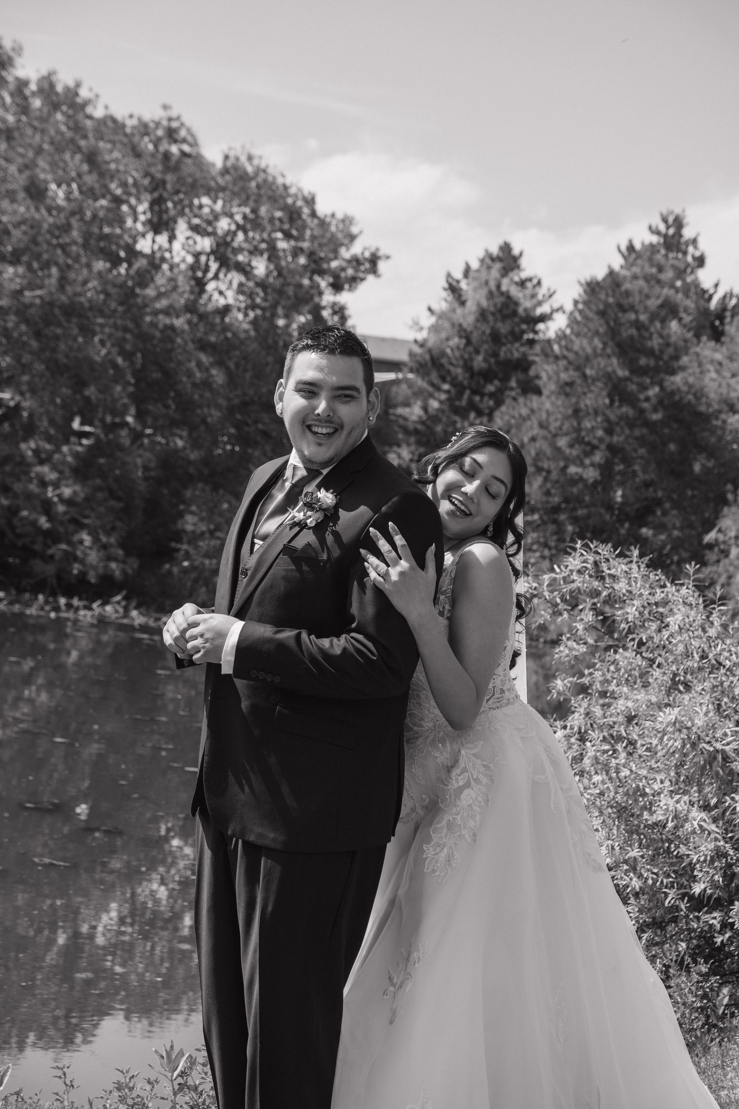 Black and white photo of a smiling bride and groom posing playfully near a reflective pond.