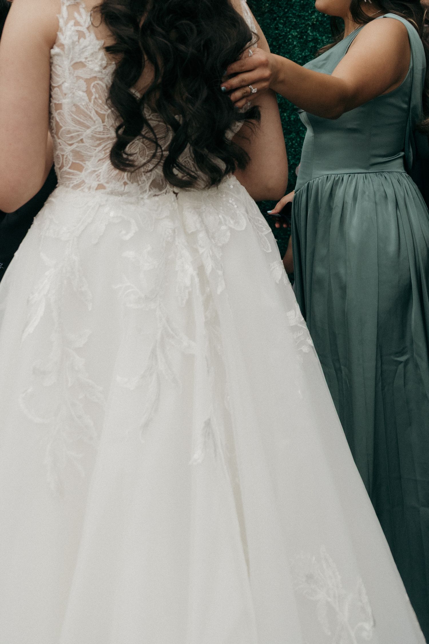 Bridesmaid in sage green dress adjusting the back of brides lace wedding gown.