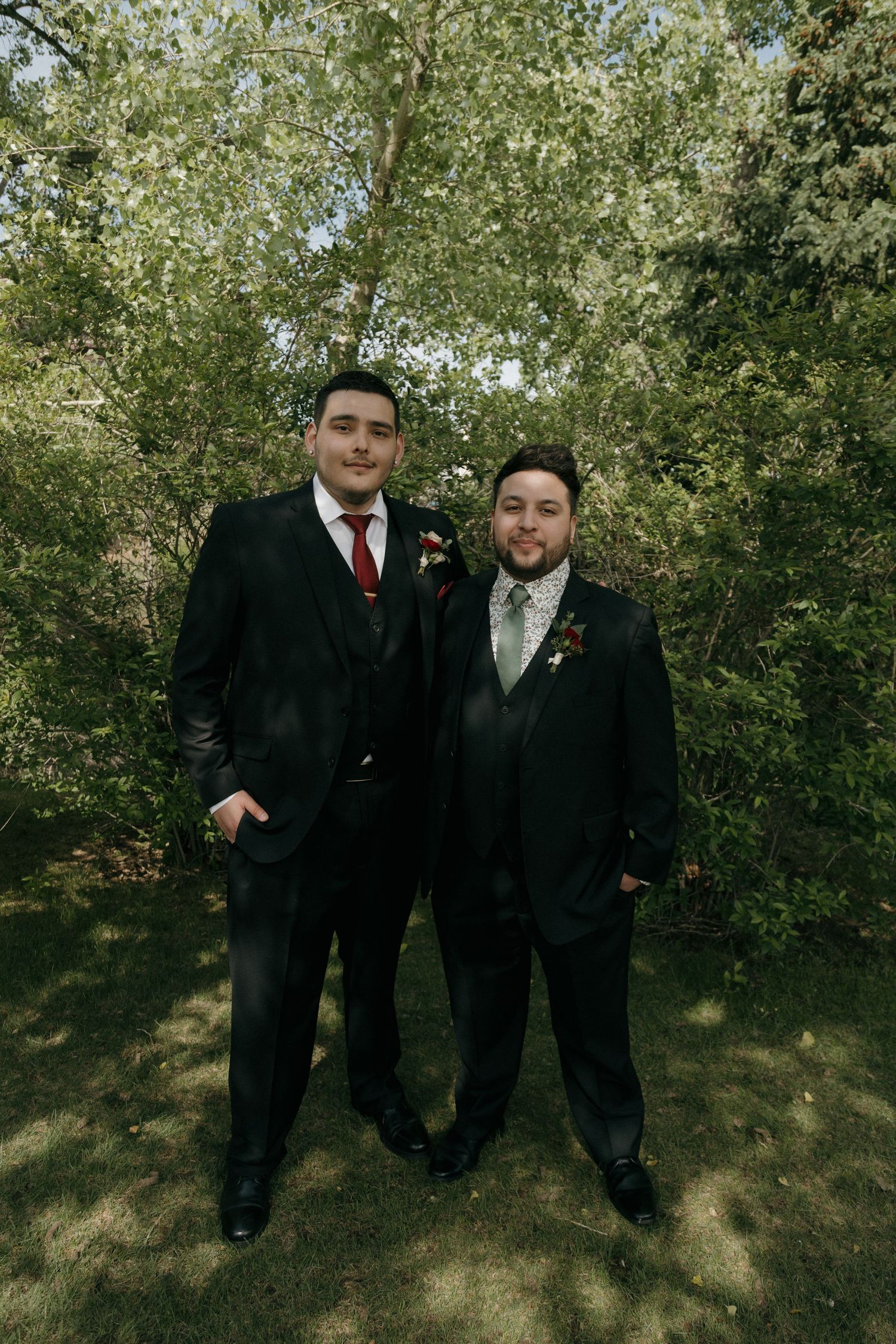 Two groomsmen in black suits with boutonnieres pose outdoors among lush green trees.