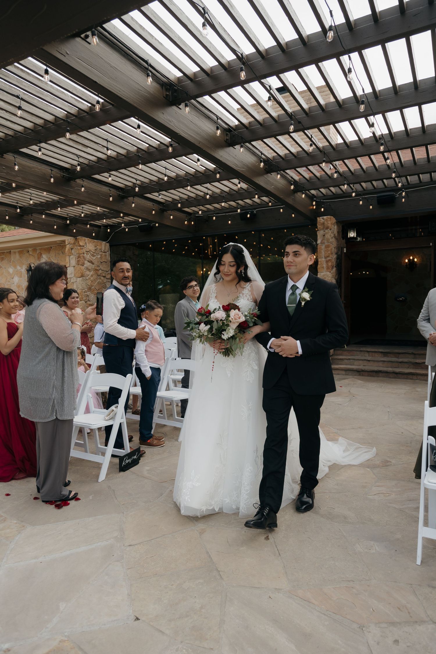 Bride in white gown and groom in black suit walk down the aisle at an elegant outdoor wedding ceremony.