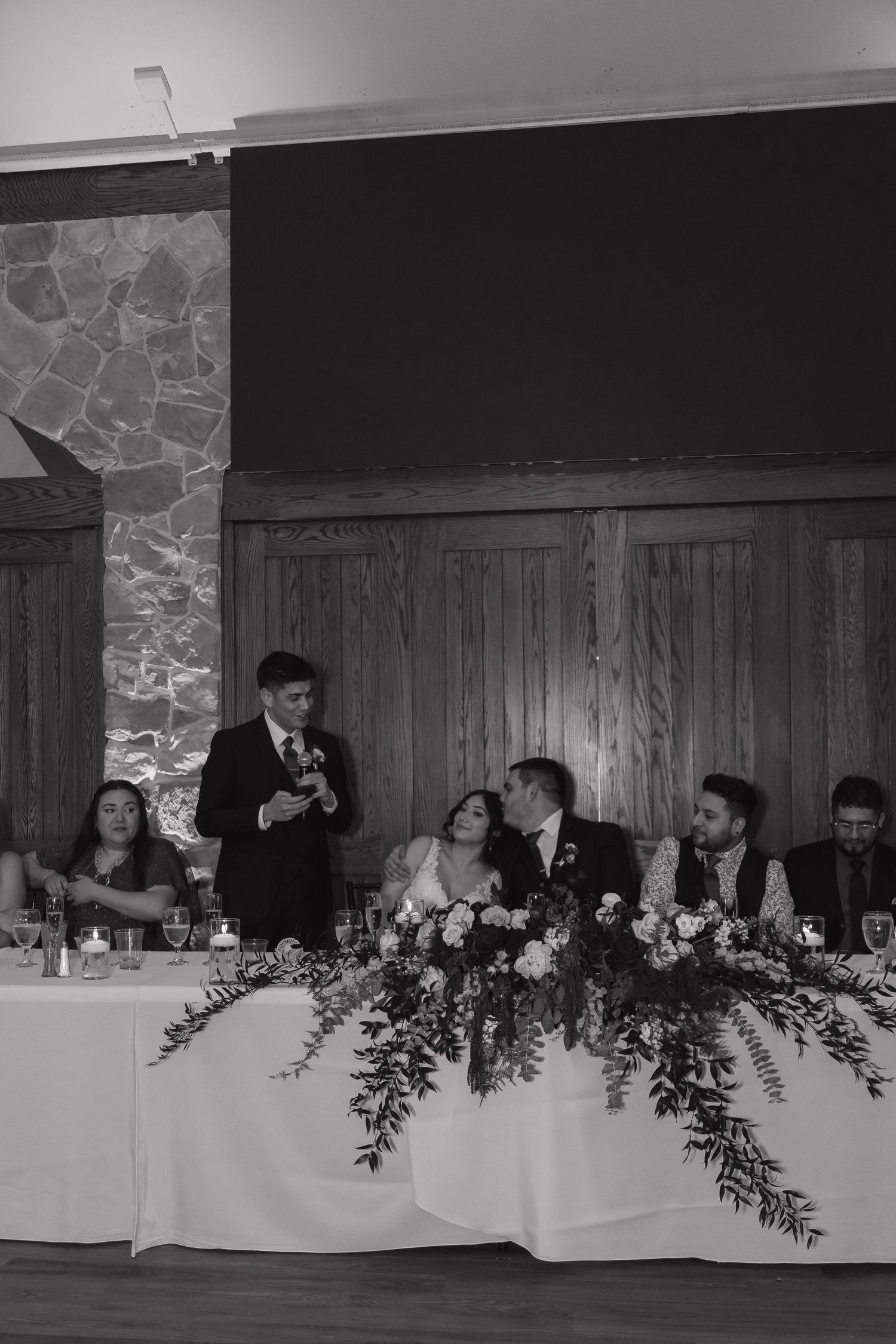 Black and white photo of wedding party at head table during reception toast with floral centerpiece.