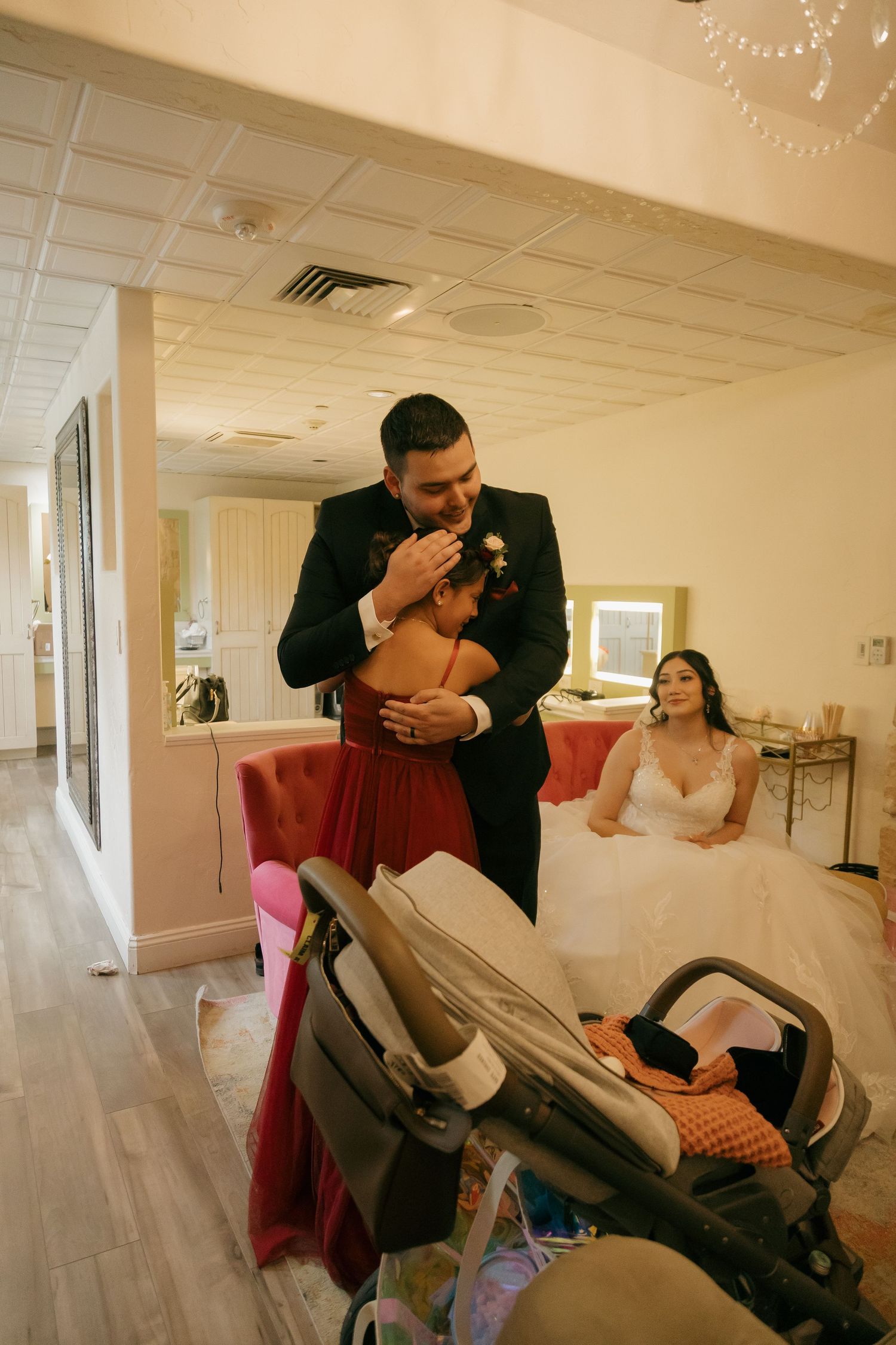 Groom in black tuxedo embracing child in red dress while bride in white gown sits in background.