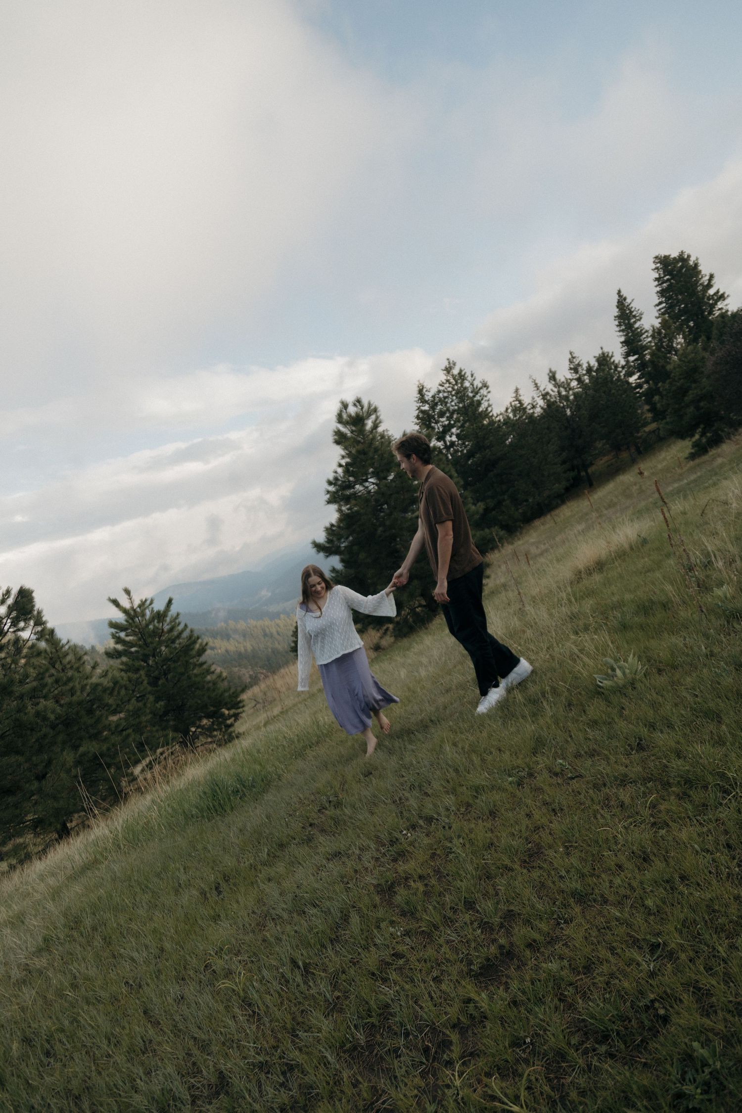 Couple holding hands while walking up a grassy hillside with mountain and pine tree views.