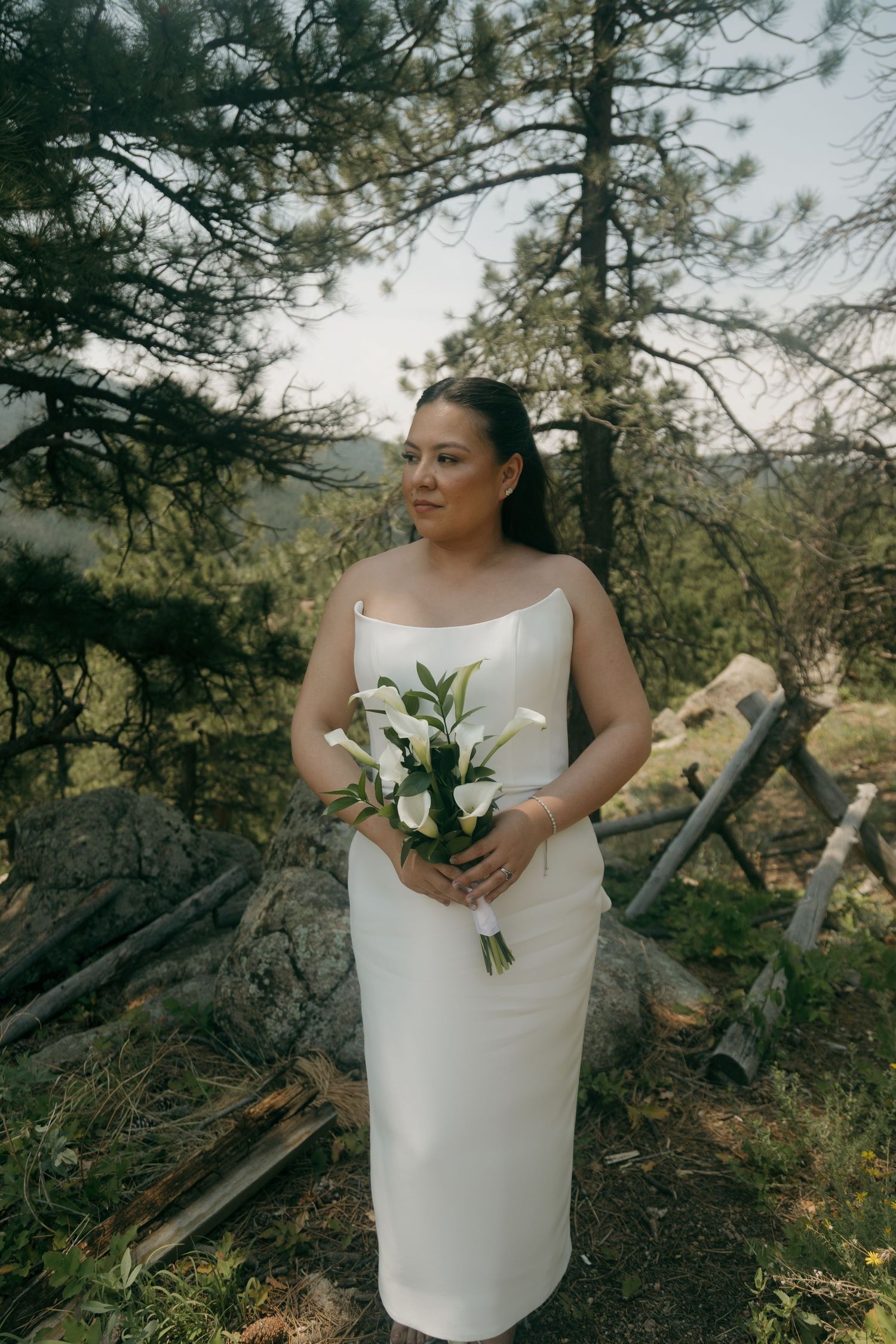A bride in a white strapless gown holds a bouquet of white calla lilies among pine trees in a mountain setting.