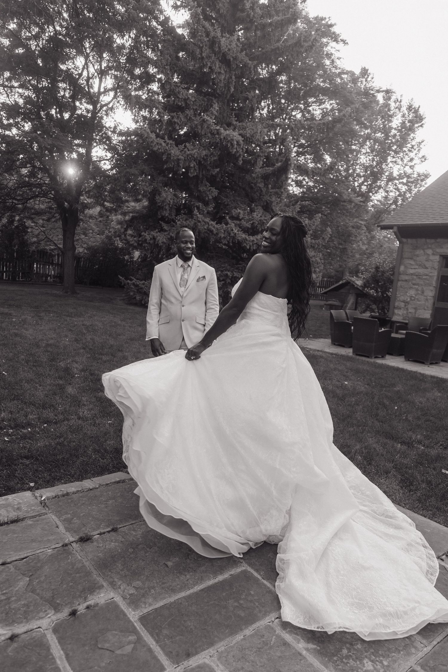 Black and white photo of Black bride twirling her ballgown while groom watches lovingly on a stone patio.