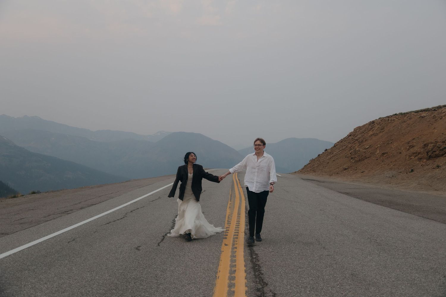 Bride and groom hold hands walking along an empty mountain highway under a hazy sky.