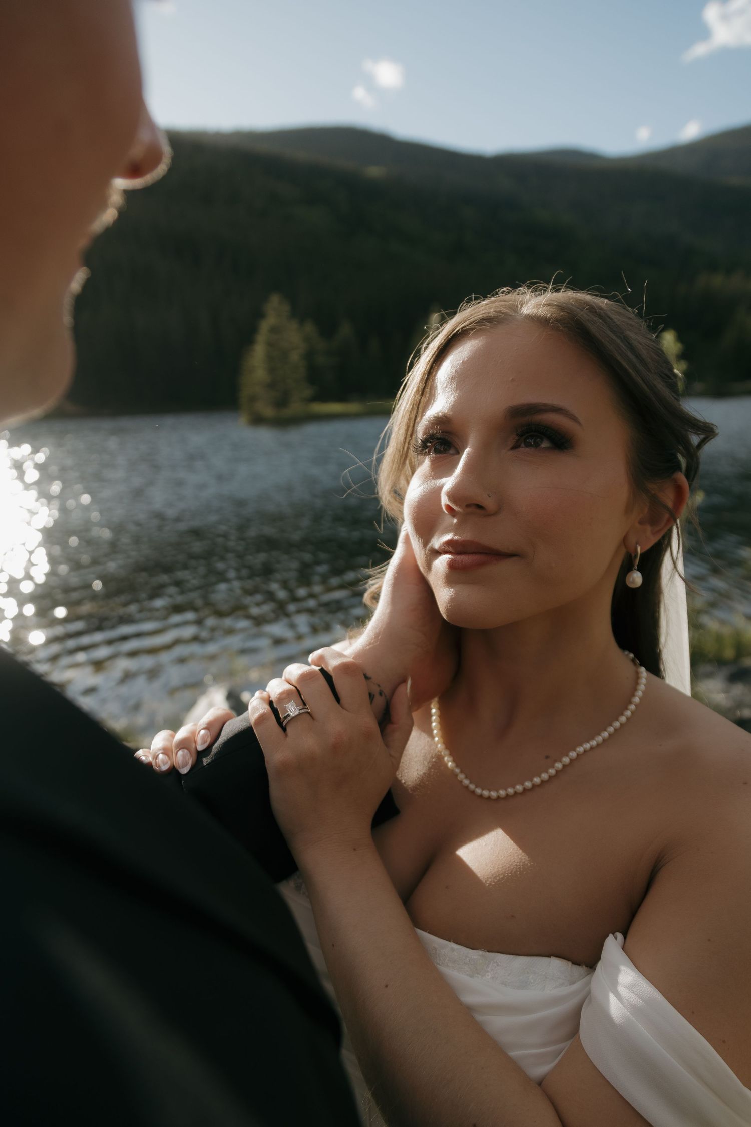Groom tenderly cups bride's face near sunlit mountain lake on their wedding day.