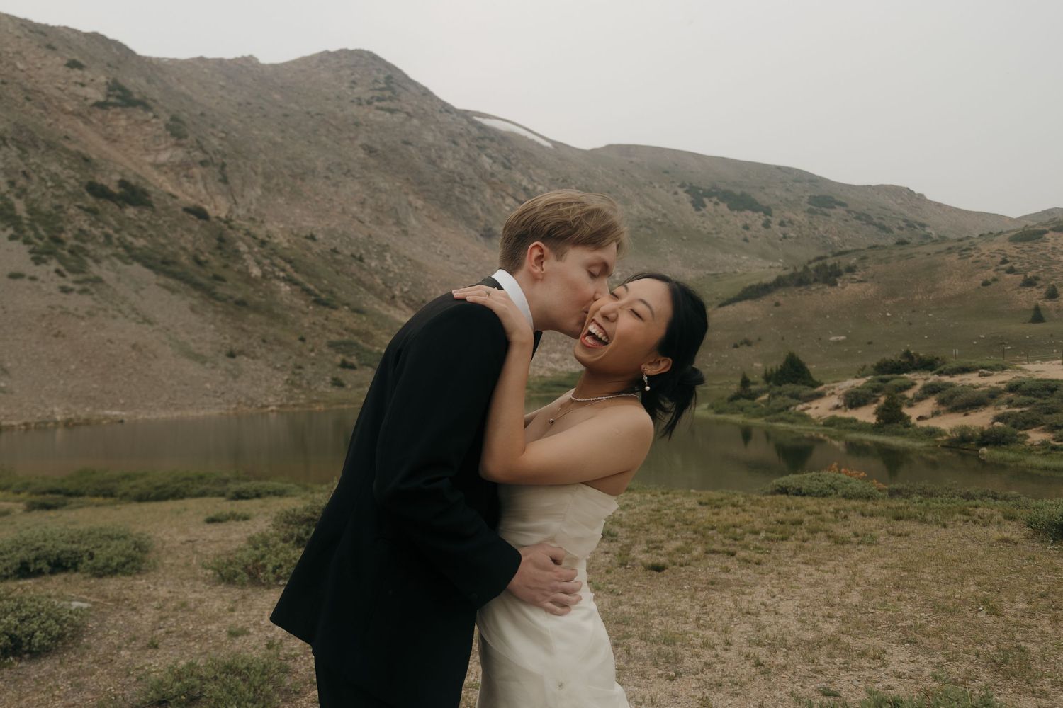 Groom kisses bride on the cheek as they embrace before a scenic alpine lake and mountain backdrop.