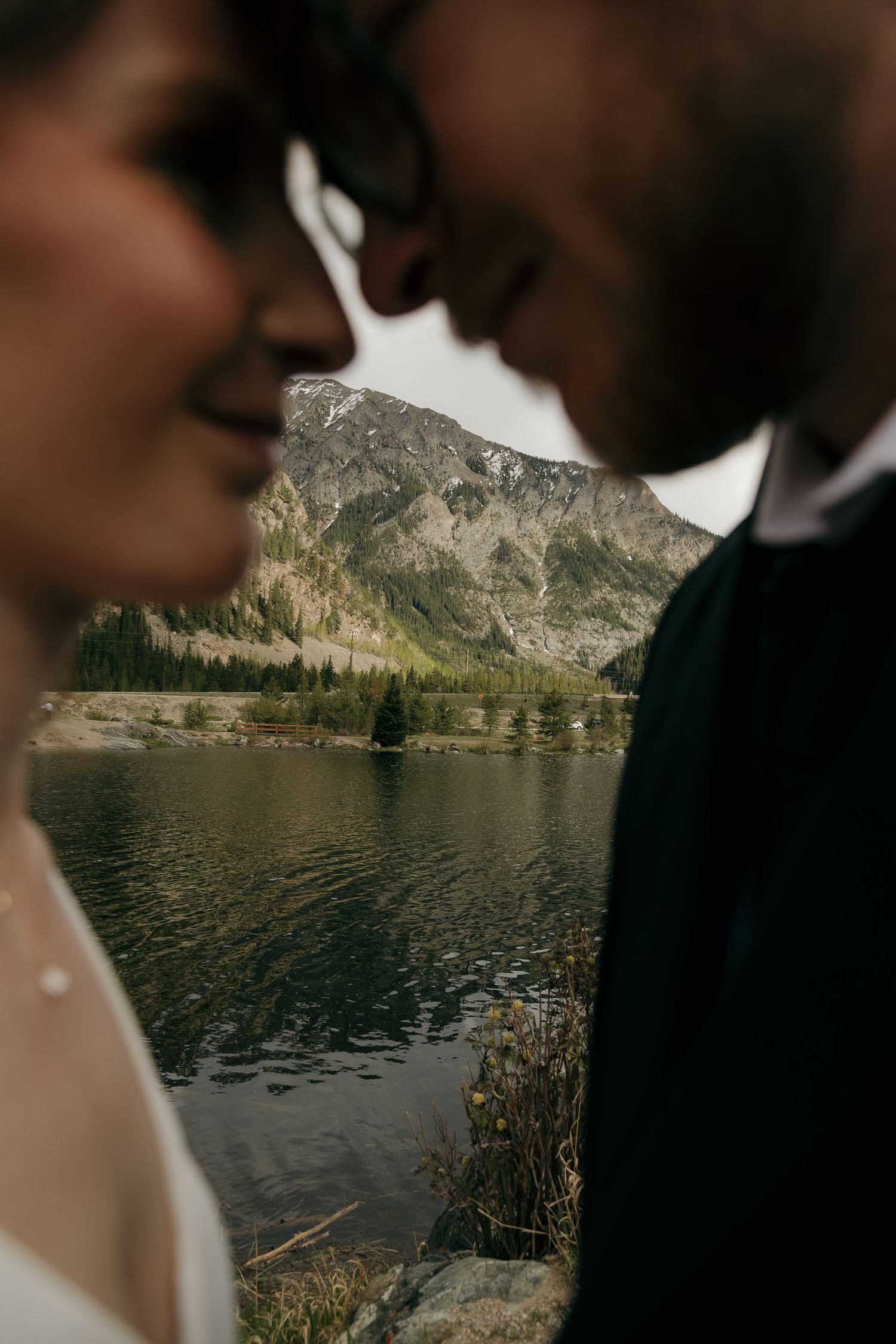 Intimate close-up of bride and groom nearly kissing with a serene mountain lake and peak in background.