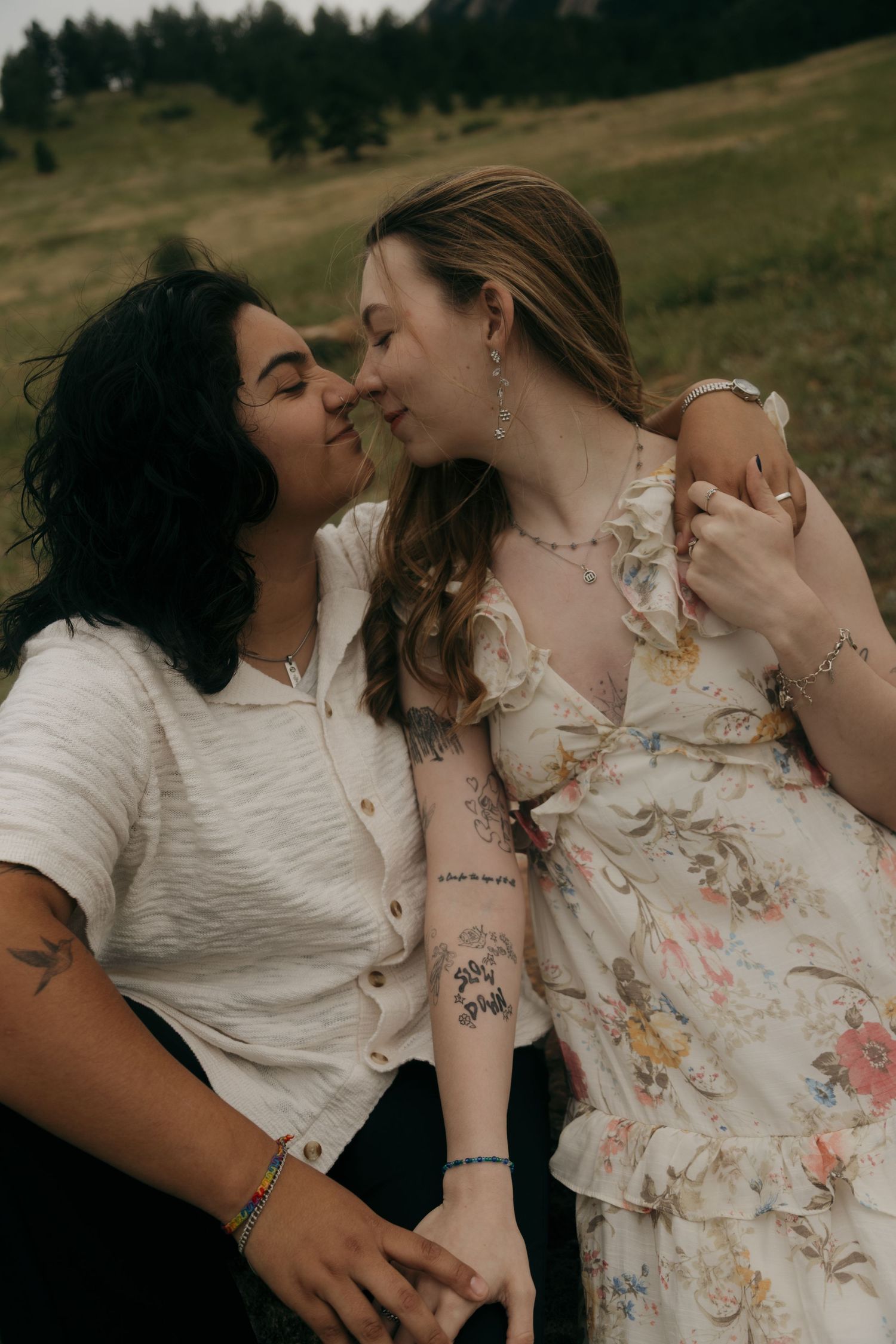 Two women with tattoos share a romantic almost-kiss outdoors in a green meadow, one in floral dress.