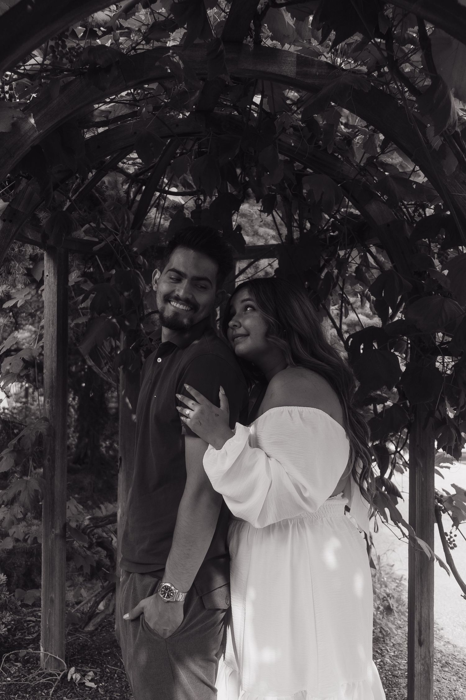 Couple embracing under a wooden garden arch with lush vines, captured in romantic black and white photography.