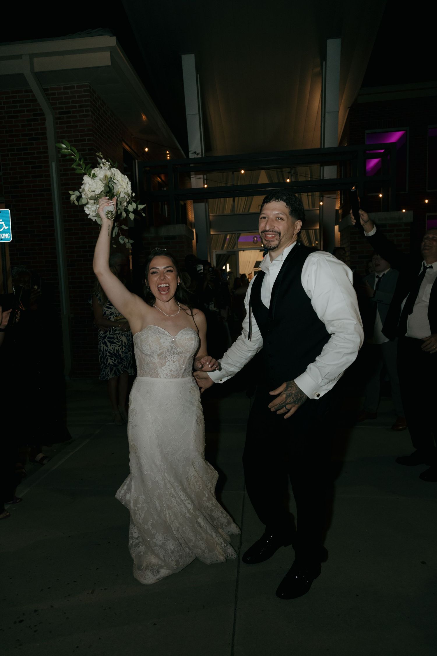 Excited bride raises bouquet while holding grooms hand during outdoor wedding reception exit.