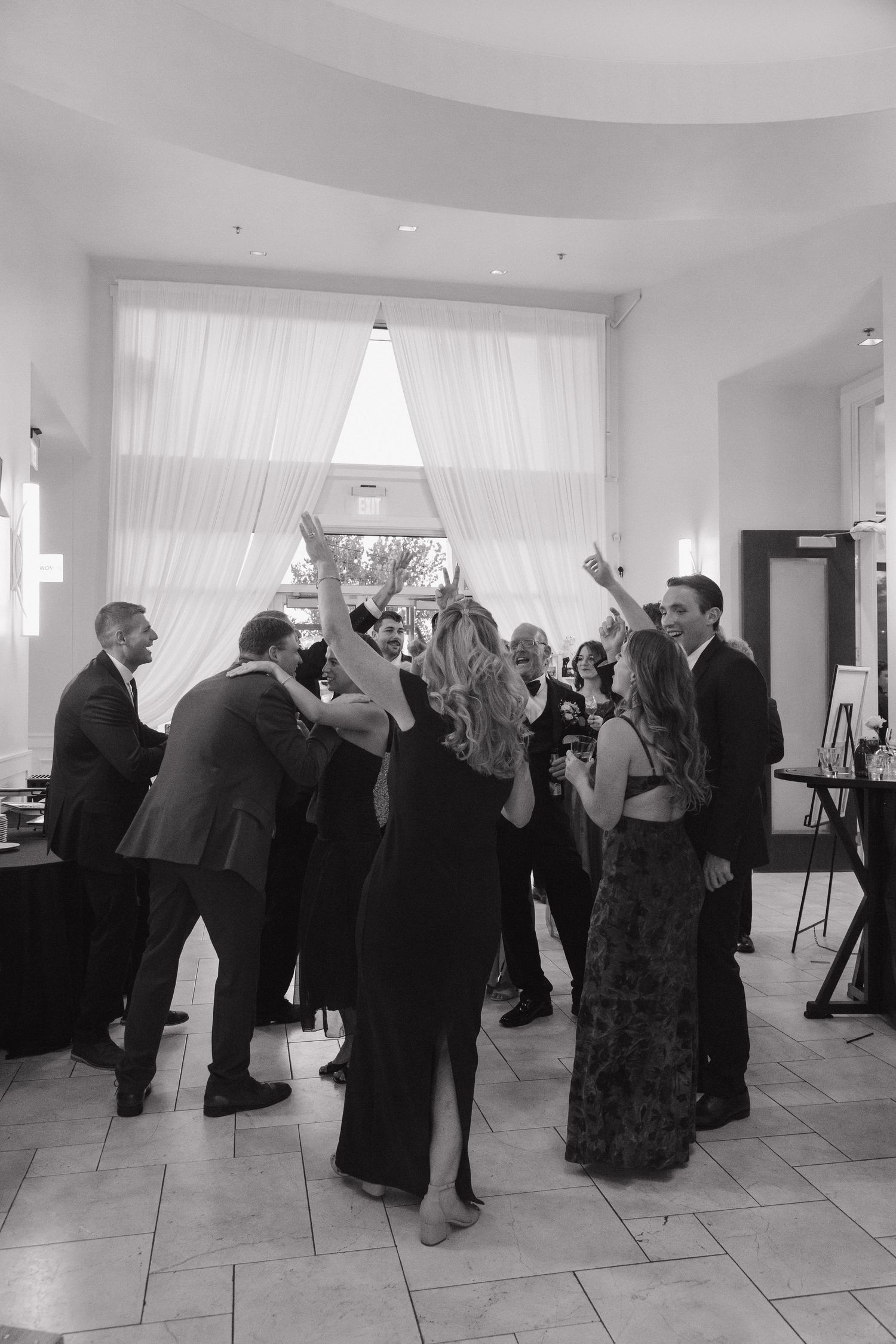 Black and white photo of wedding guests dancing and celebrating at elegant indoor reception.