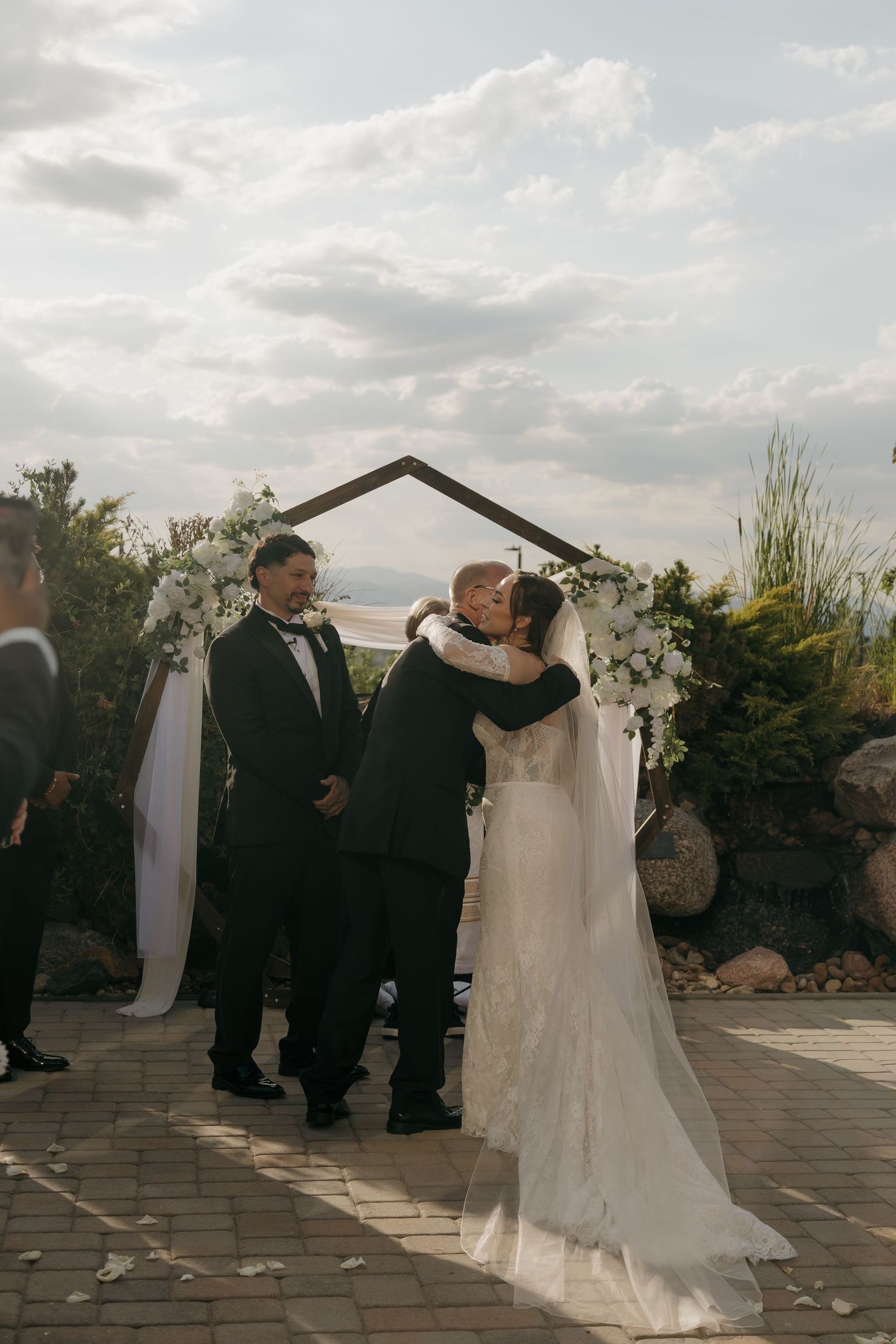 Bride and groom share first kiss under floral arch at outdoor wedding ceremony with scenic sky.