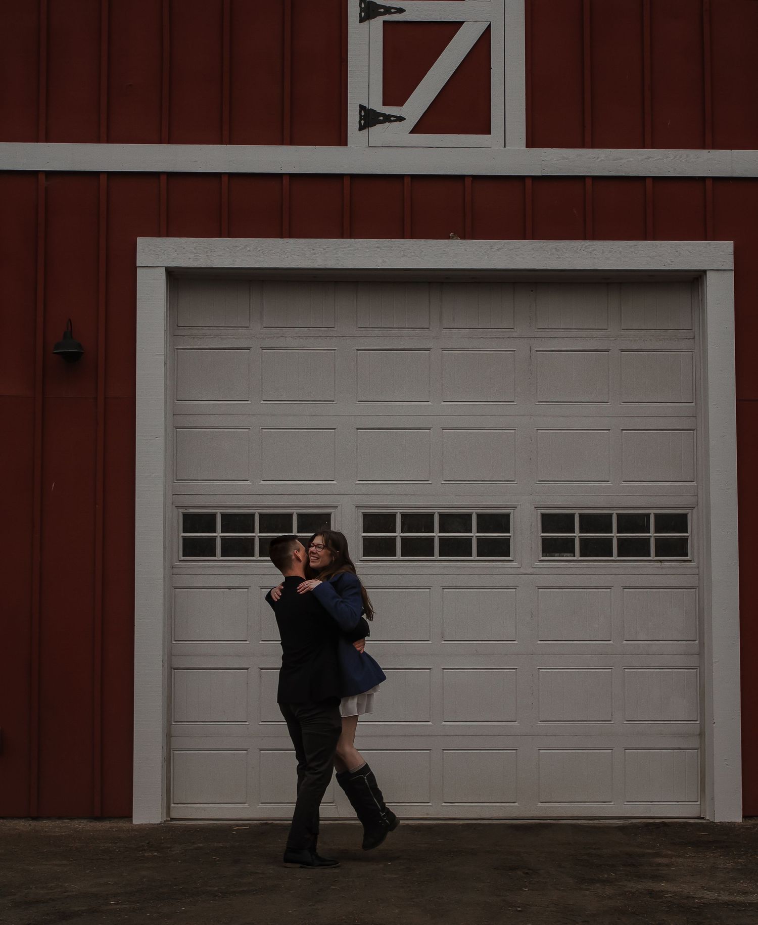 A couple embracing against a white garage door on a red barn.