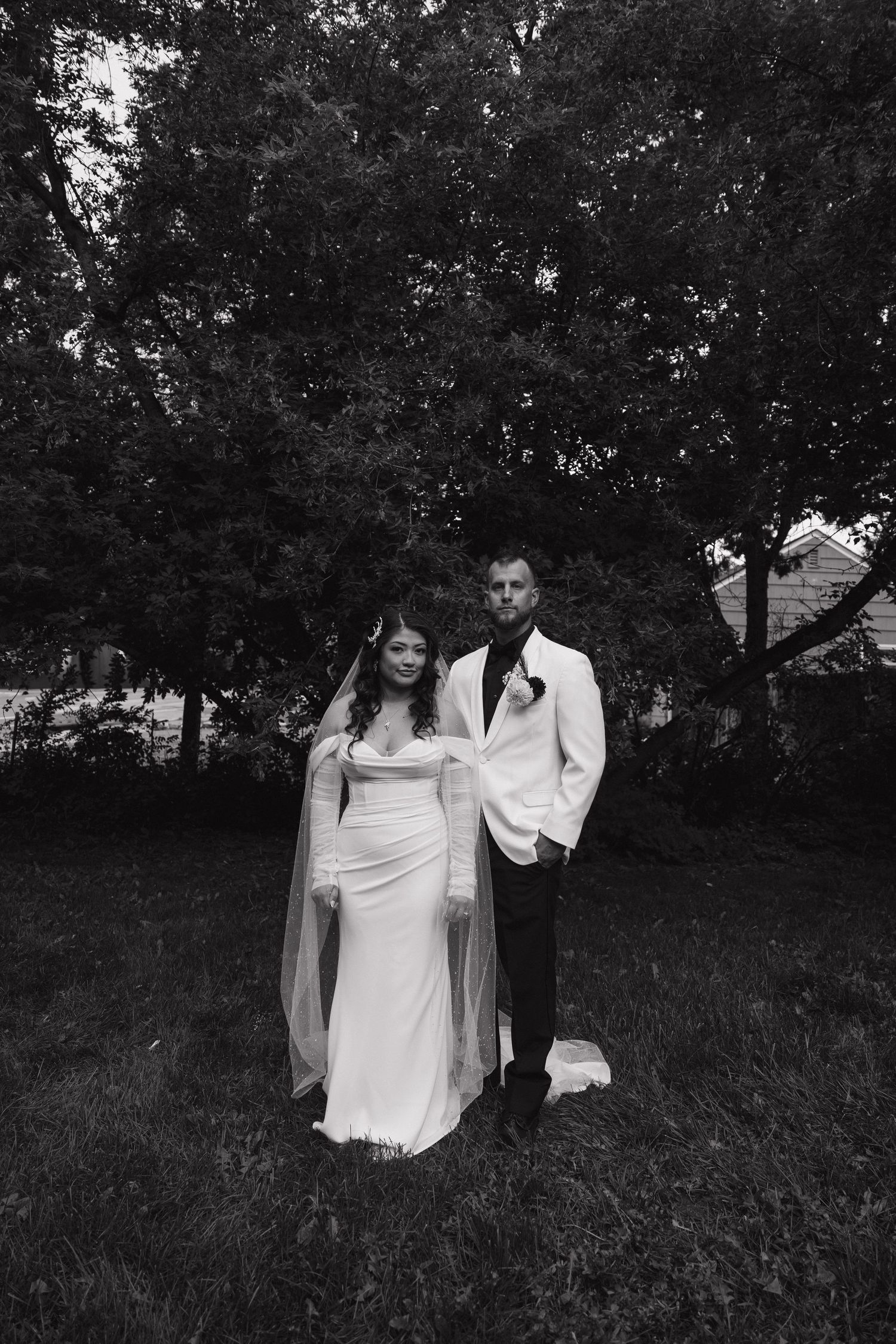 Black and white portrait of bride and groom standing together under large trees in garden setting.