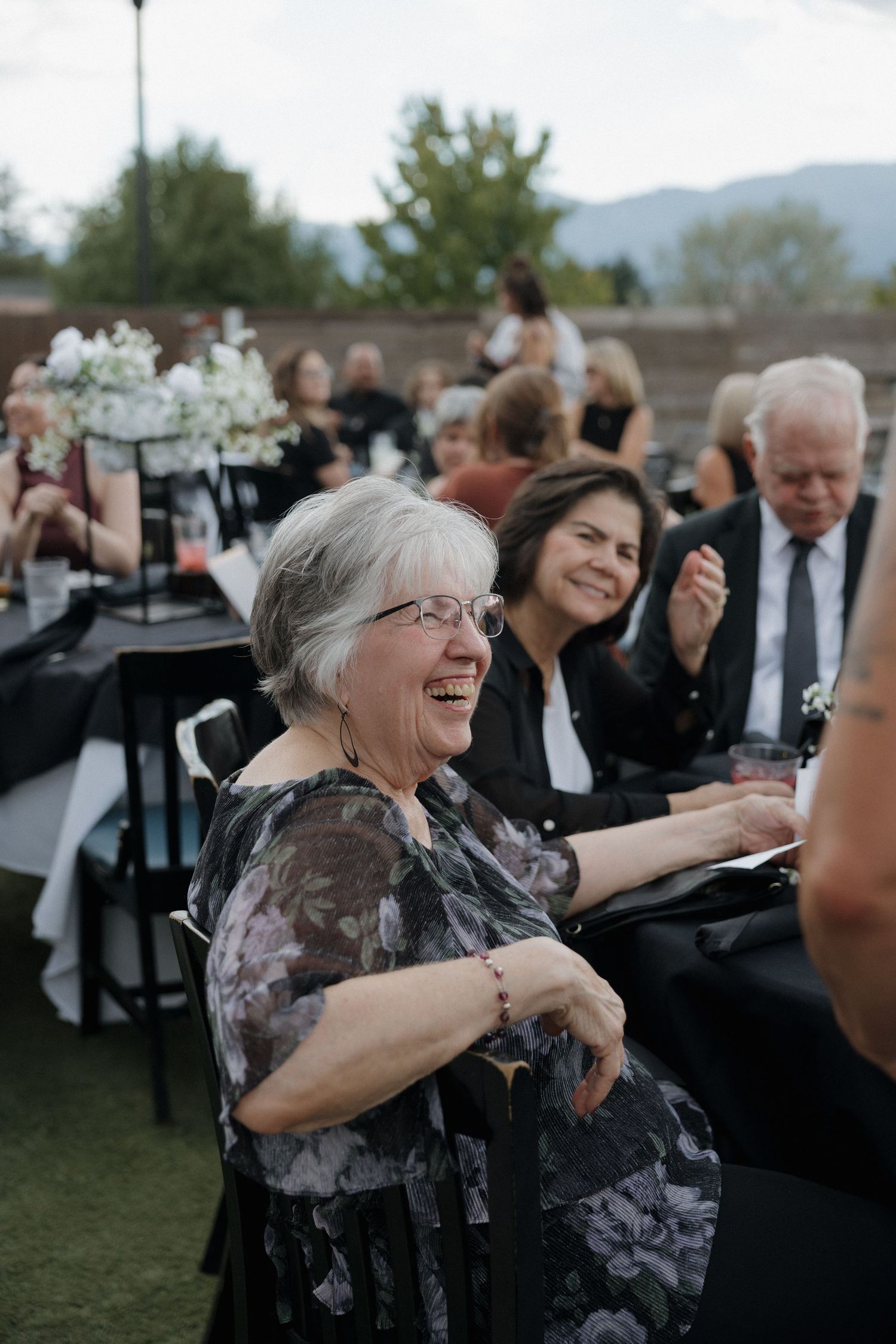 Elderly woman laughing joyfully at outdoor wedding reception with guests in background.