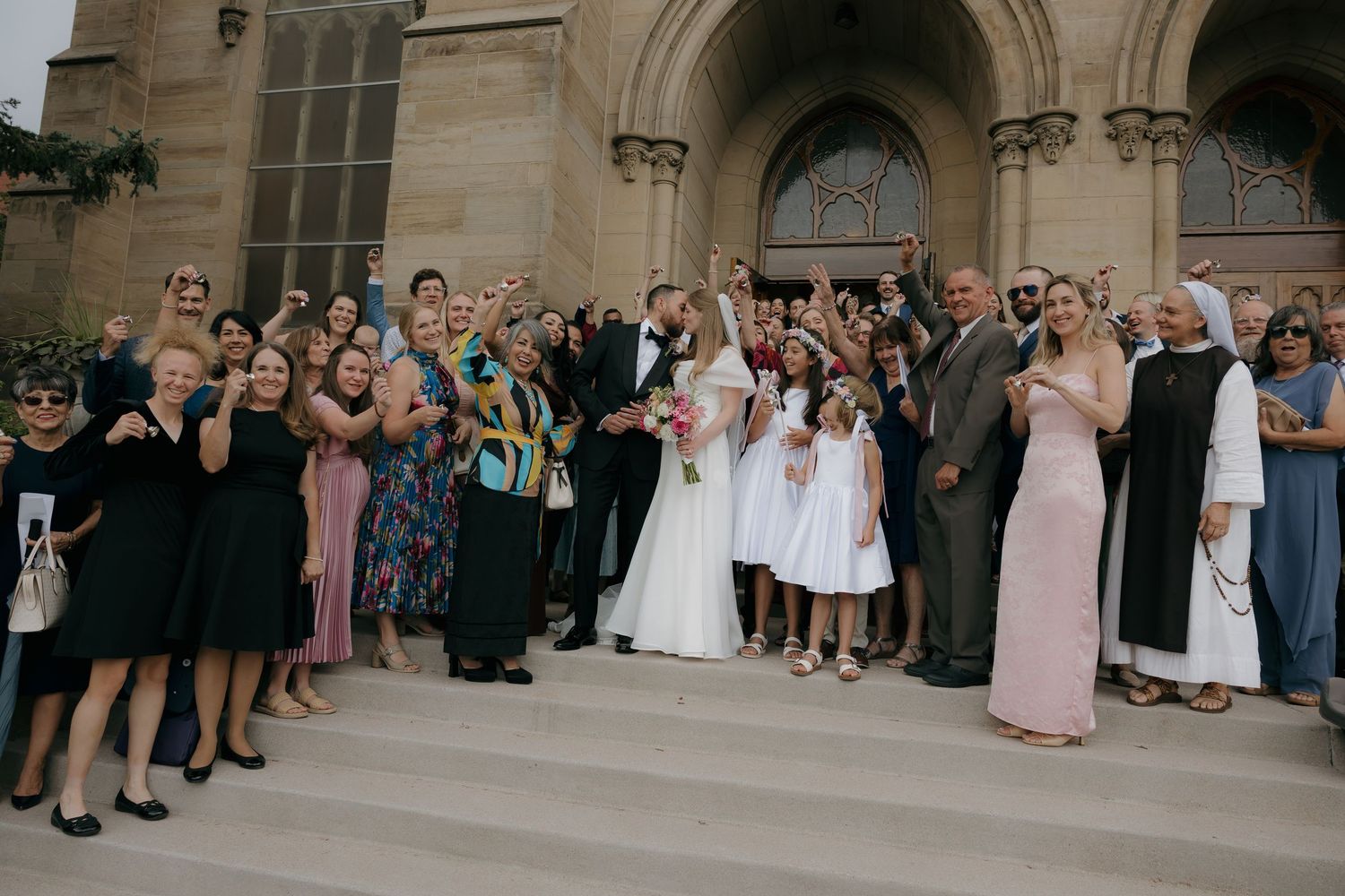 Wedding party celebrating on church steps with bride and groom kissing surrounded by cheering guests.