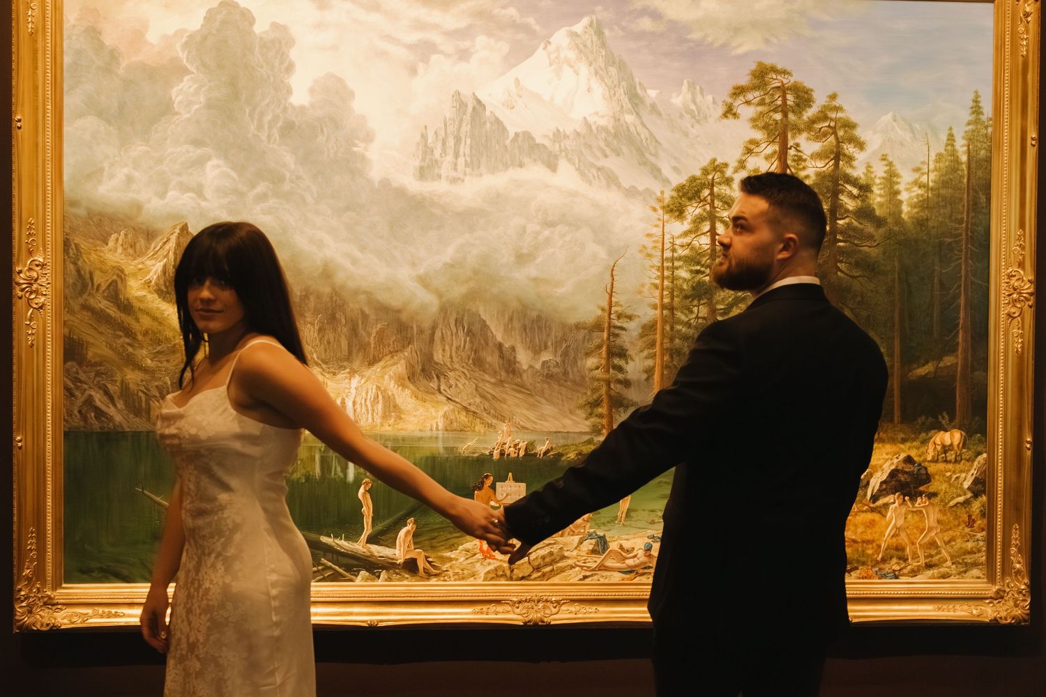 Art gallery visitors admire paintings of mountainous landscapes displayed in ornate gold frames on a museum wall.