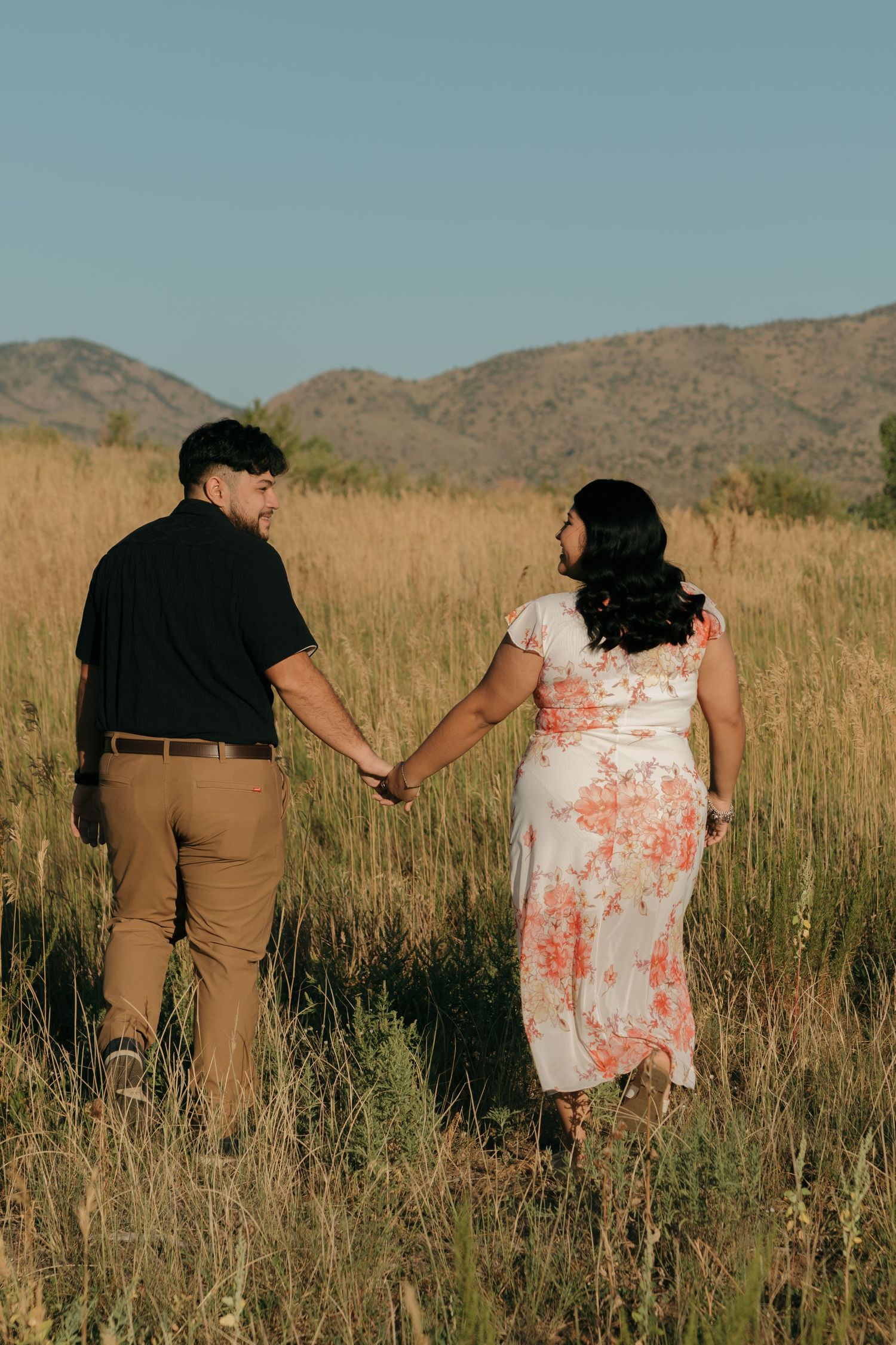 A couple holds hands while walking through tall grass with mountains in the background.