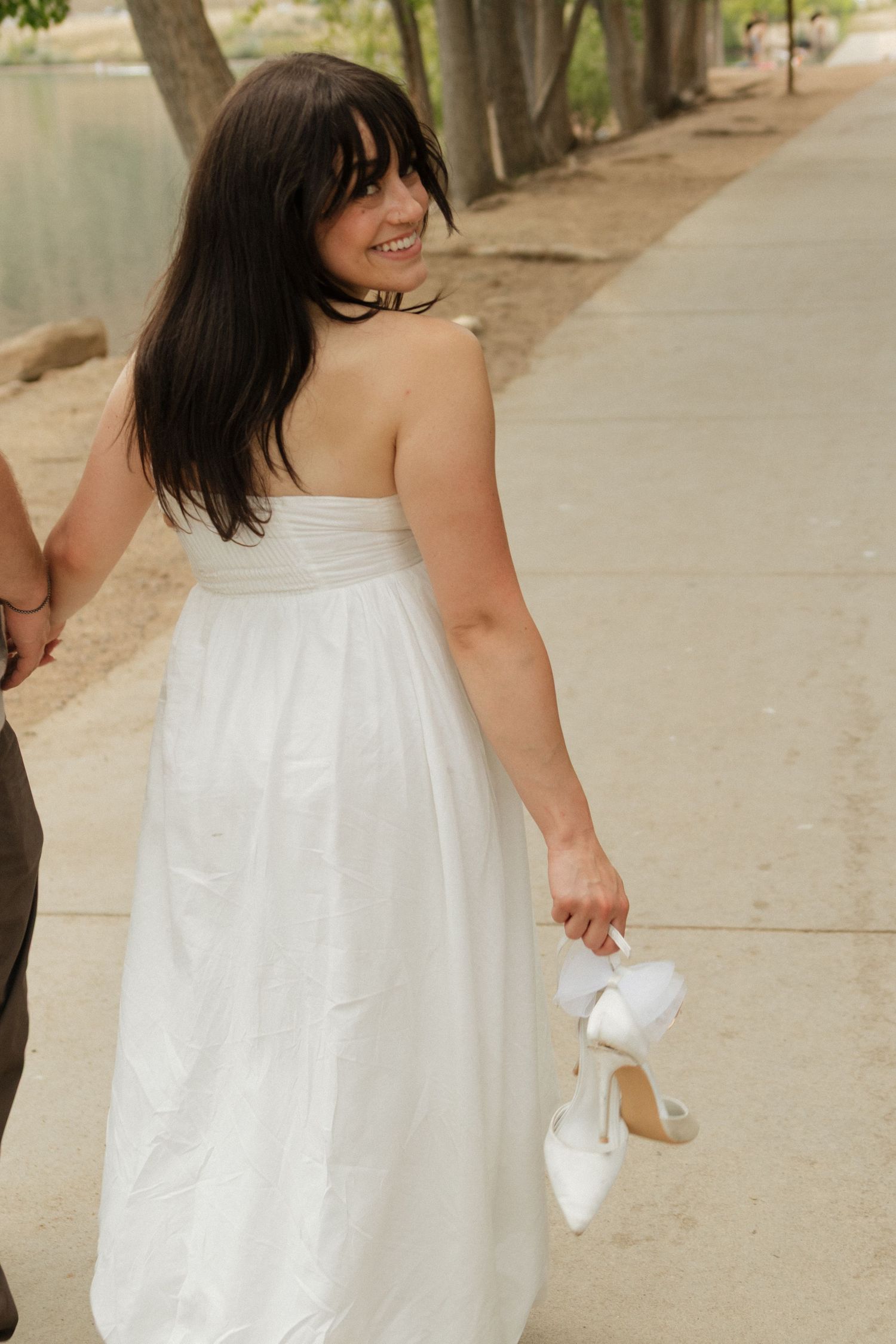 Woman in white summer dress walks down a sunny path while holding an elegant purse.