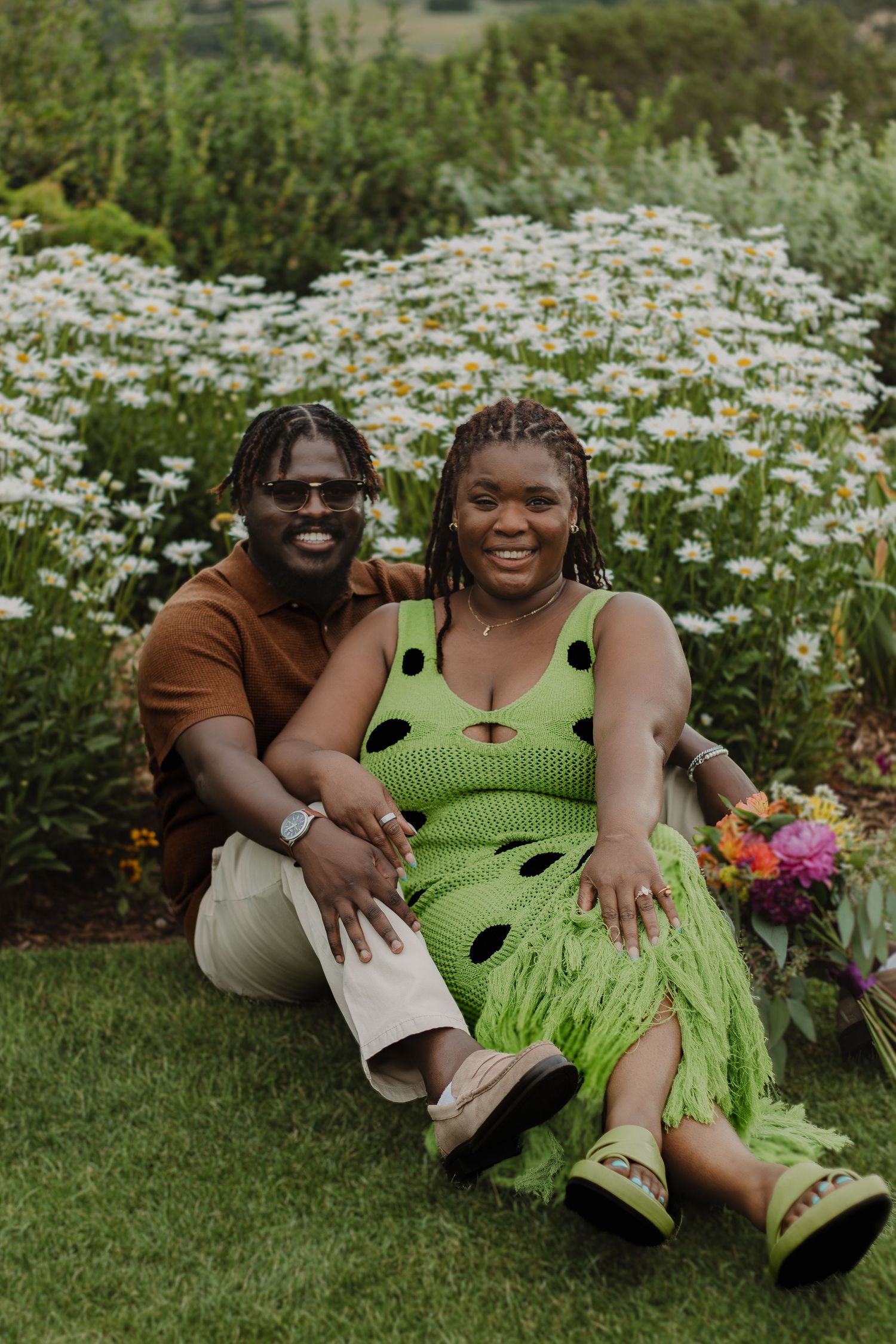 A couple sits together on grass surrounded by blooming daisies, with the person wearing a lime green polka dot dress.