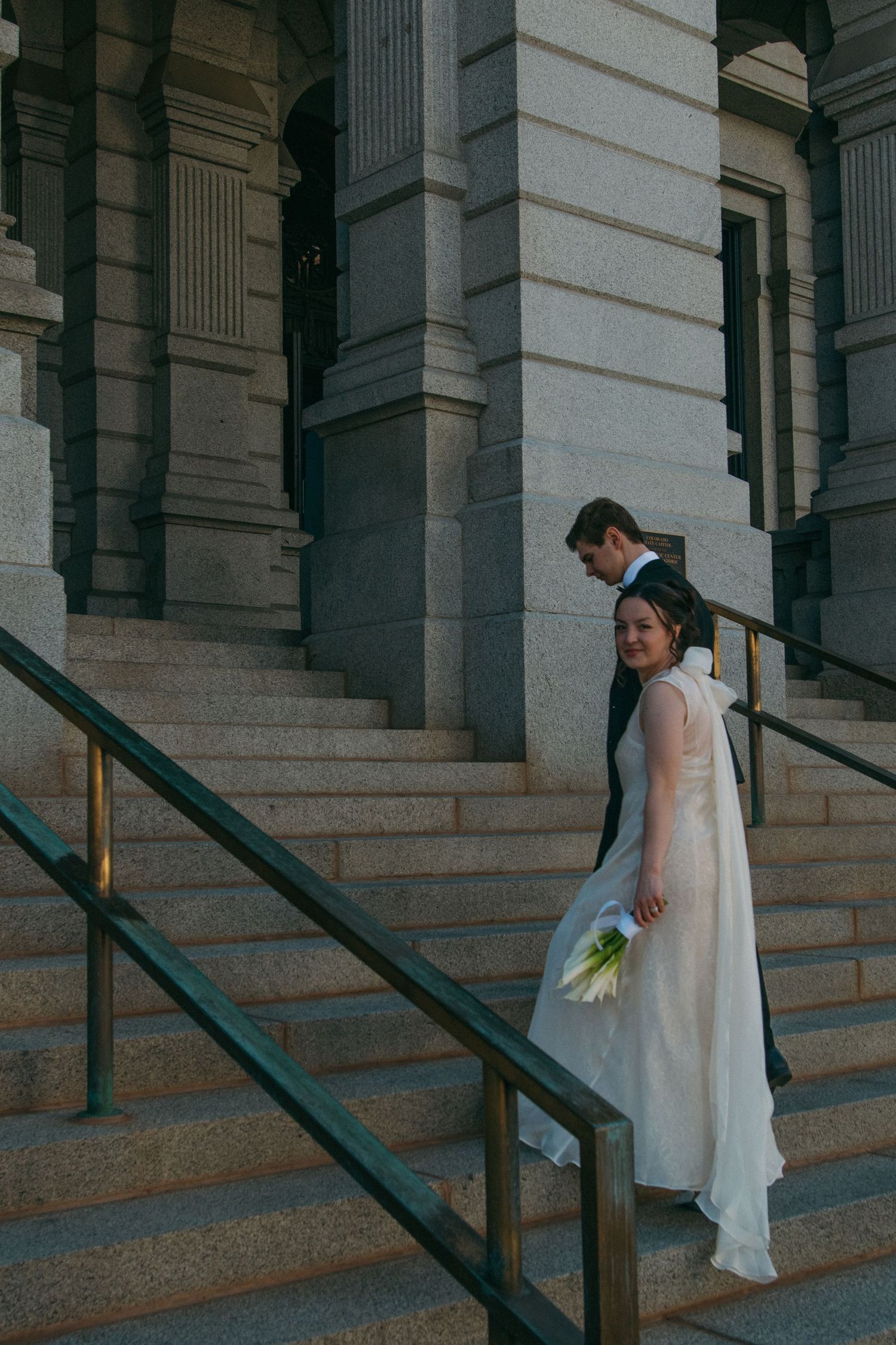 A couple in wedding attire descends stone steps of a classical building.