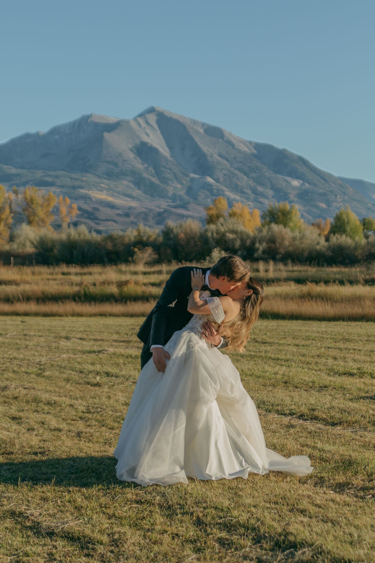 Newlyweds share an intimate moment in a field with majestic mountains and autumn colors in the background.