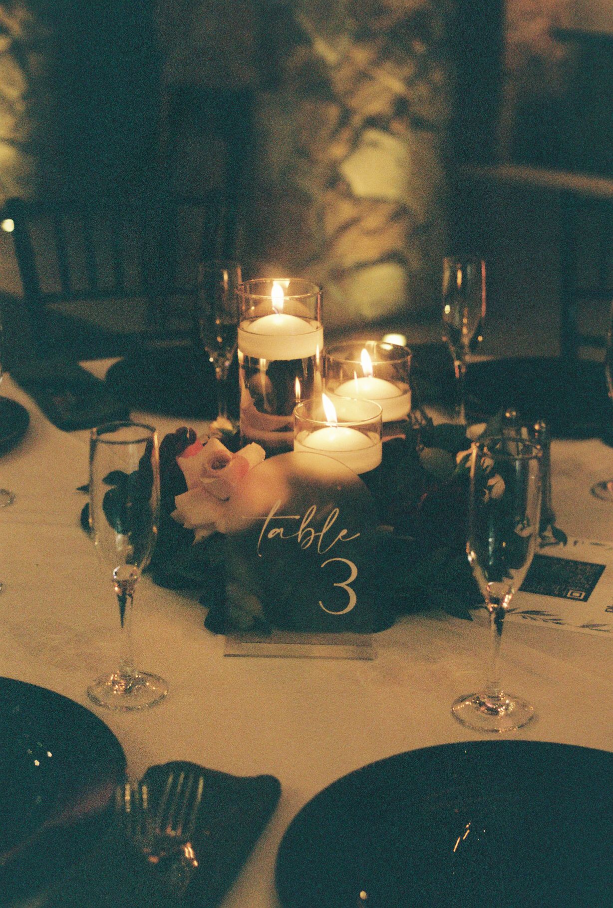 Elegant wedding table 3 centerpiece with floating candles, pink roses, dark foliage, and crystal glassware in moody lighting.
