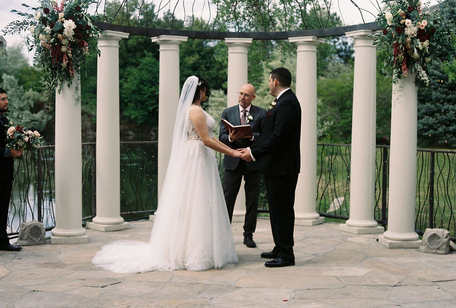 Bride and groom exchange vows at outdoor wedding ceremony under elegant white columns with floral arch decorations.