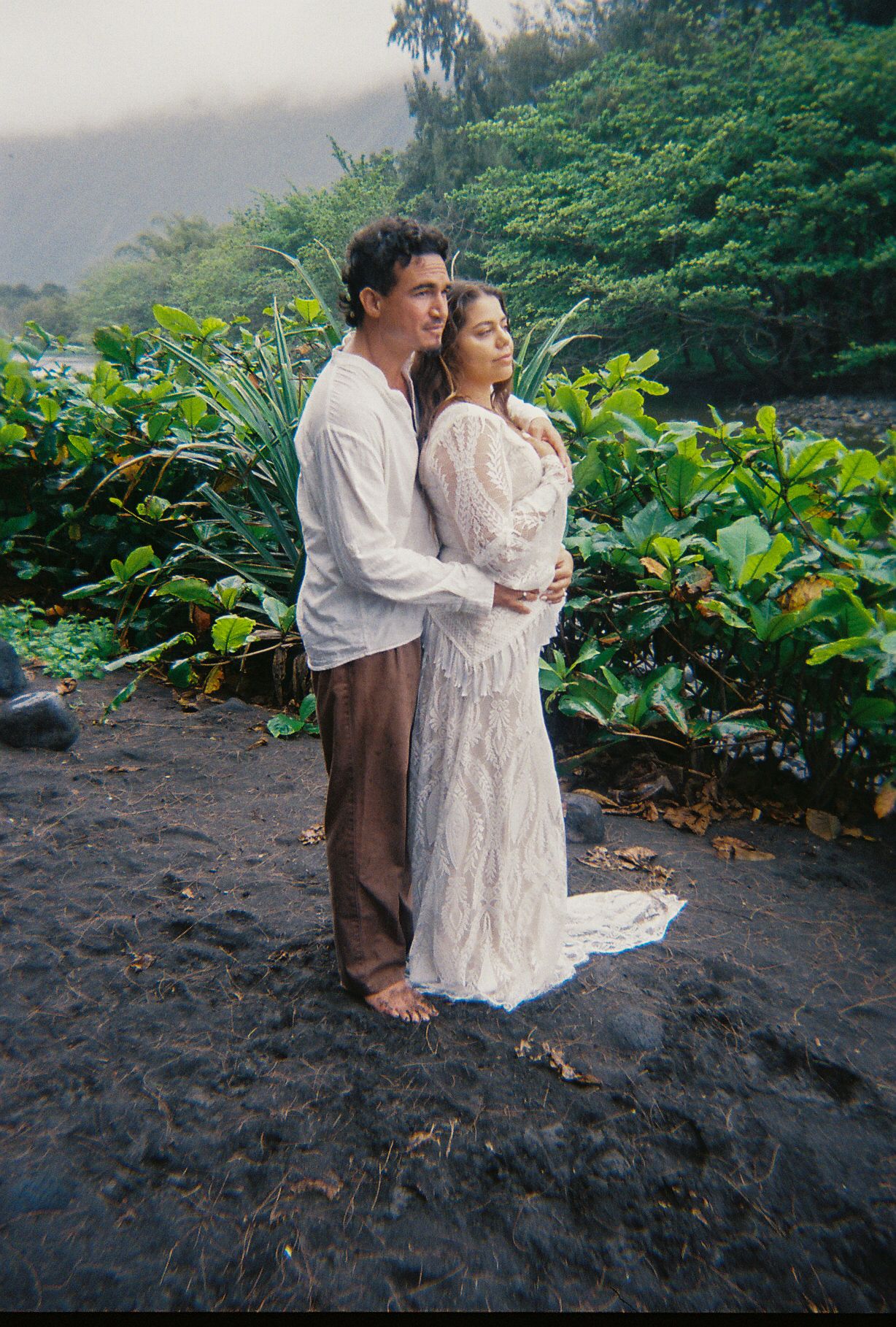 Couple embracing on volcanic black sand surrounded by lush tropical foliage on film photograph.