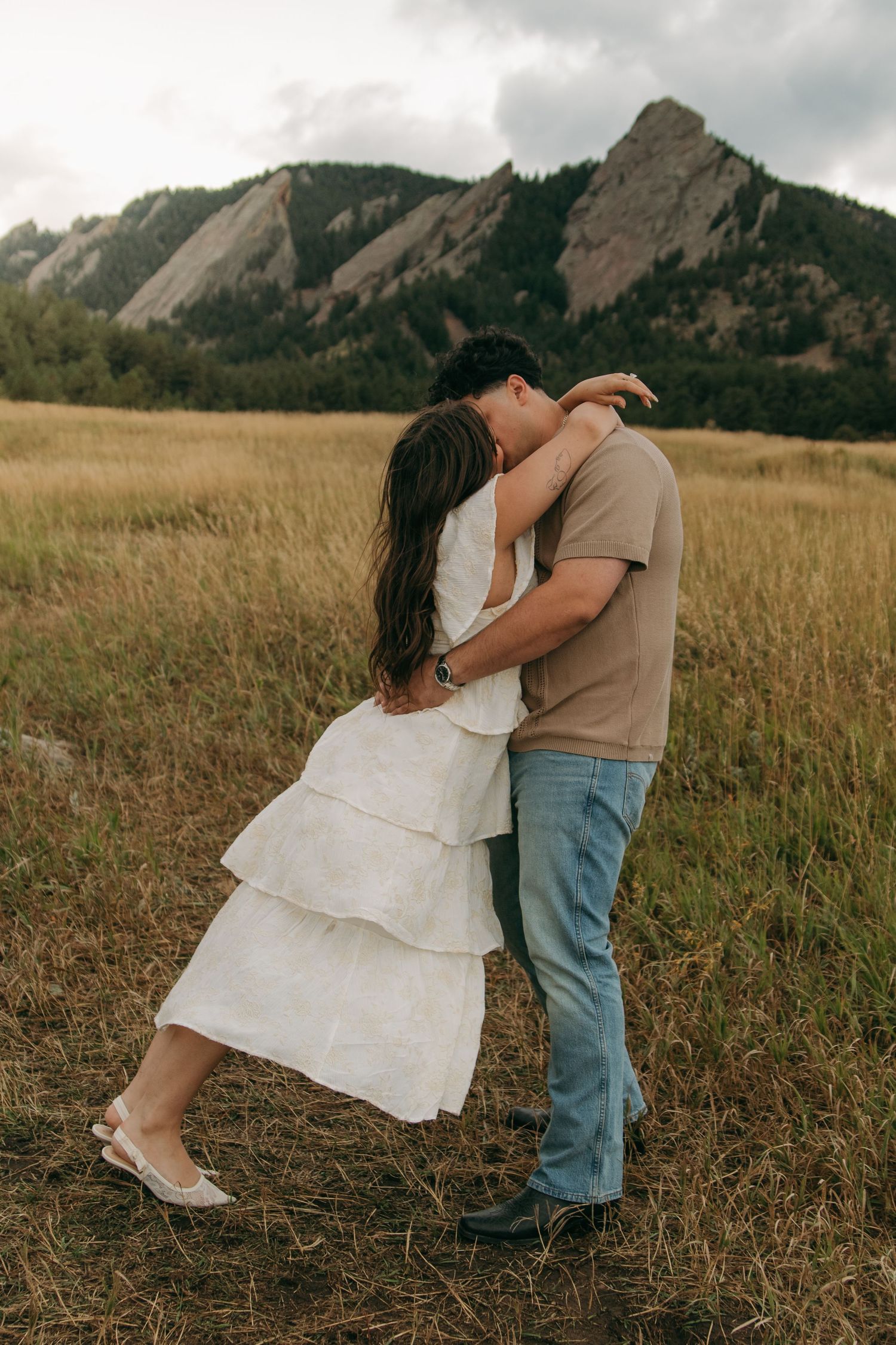 Engaged couple shares tender embrace on dirt path in golden grass field with Flatirons mountains backdrop.