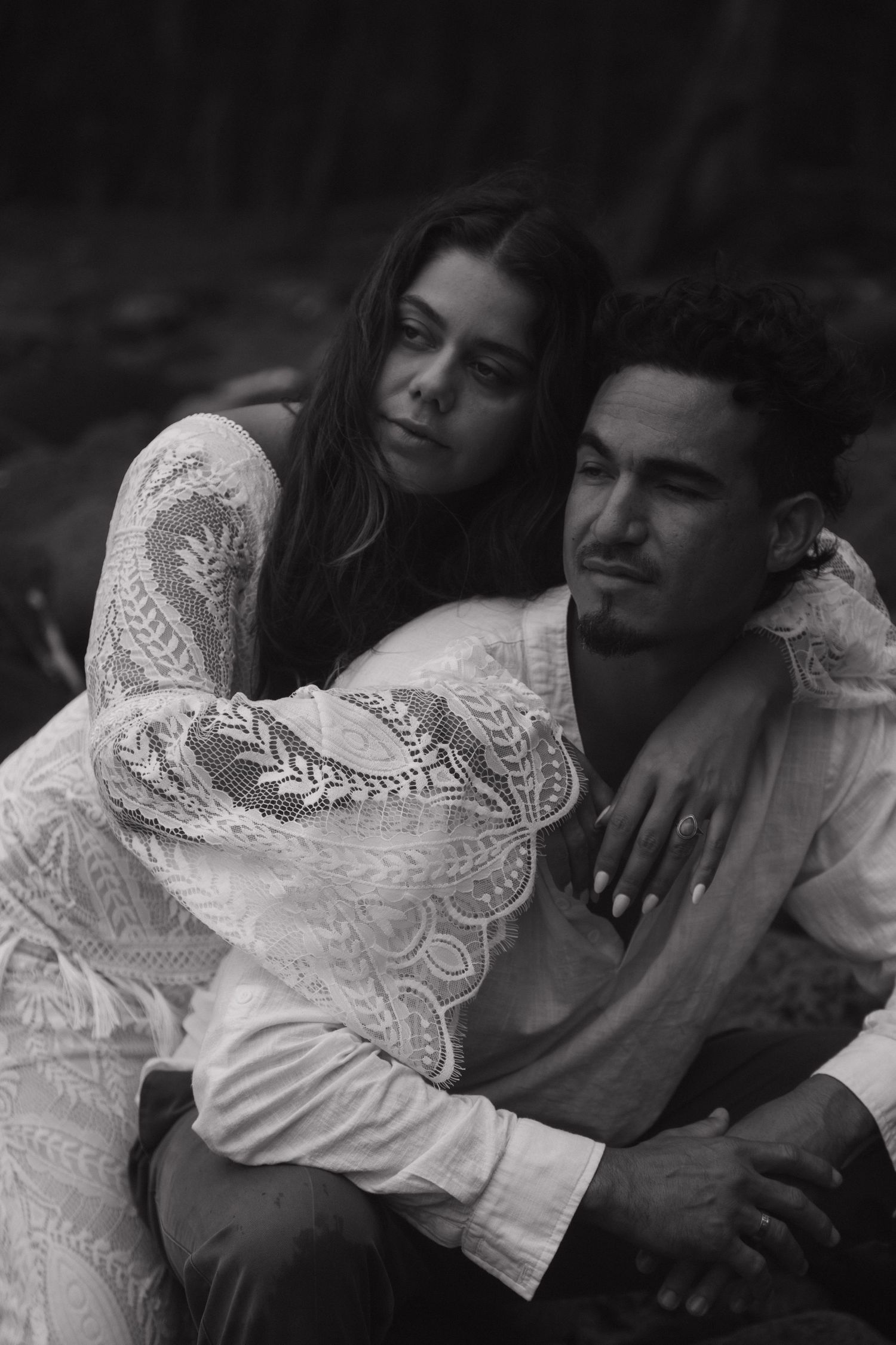 Black and white portrait of couple sitting together outdoors, woman in lace dress embracing man from behind.