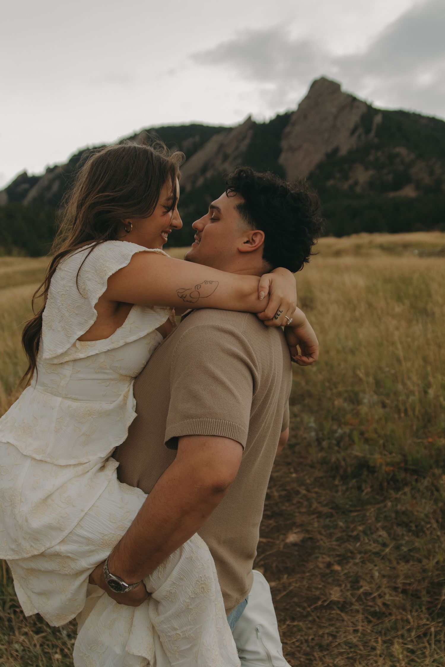 Man lifting smiling woman in white dress in a field with the Flatirons mountains in the background.