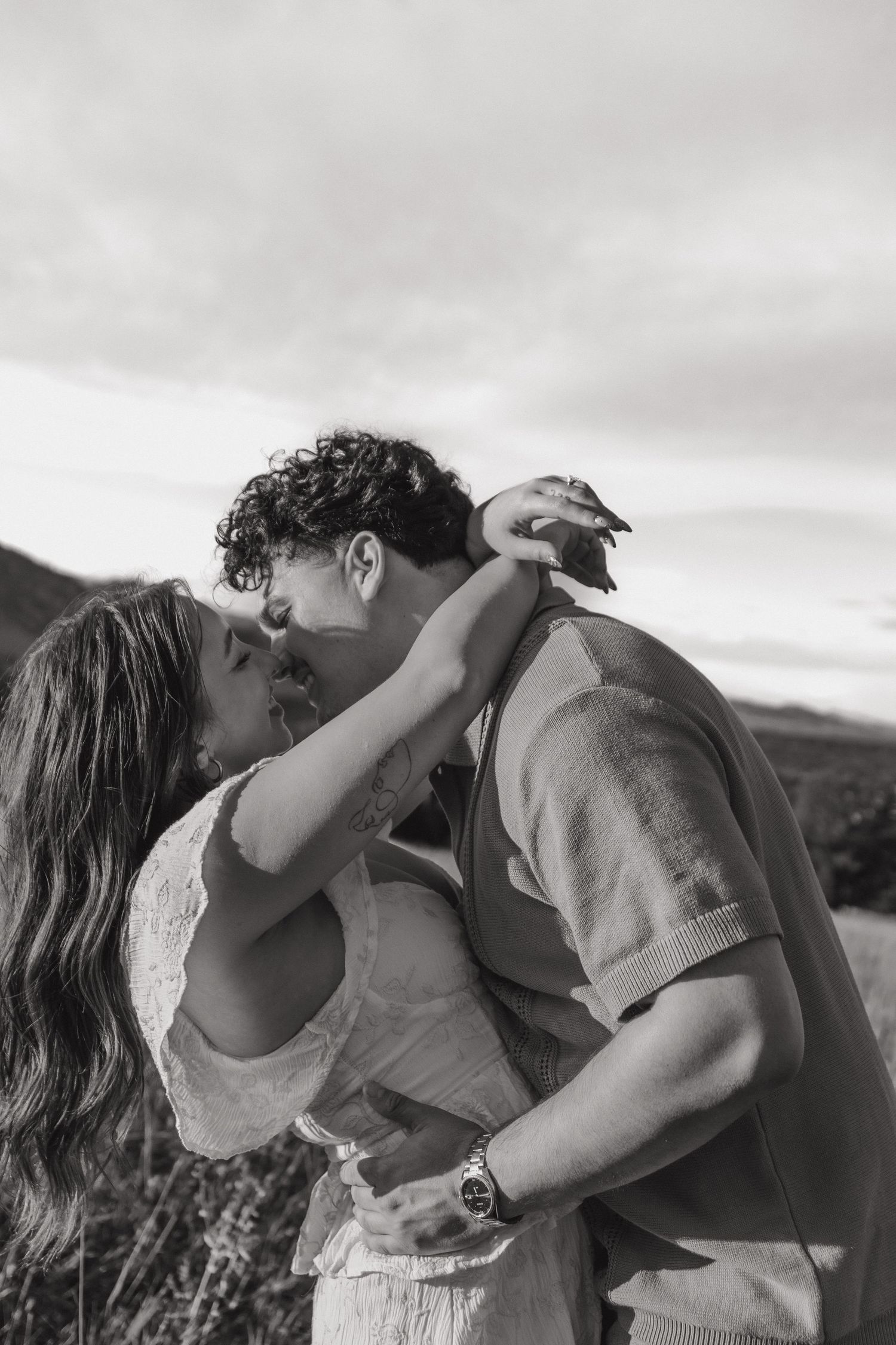 Black and white photo of a romantic couple sharing a kiss outdoors with mountains in the background.