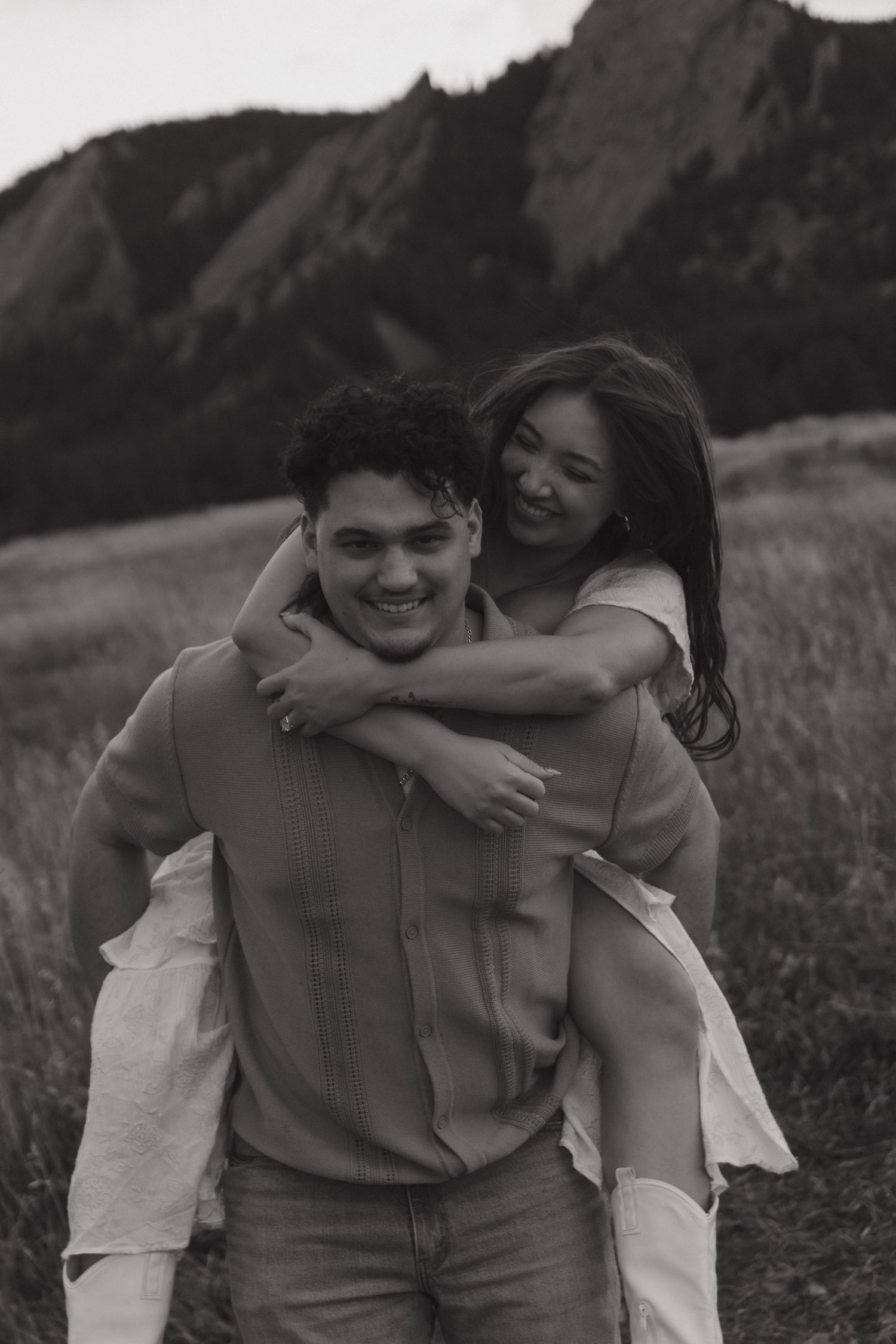 Black and white photo of a smiling couple, man giving woman a piggyback ride in a mountain meadow.