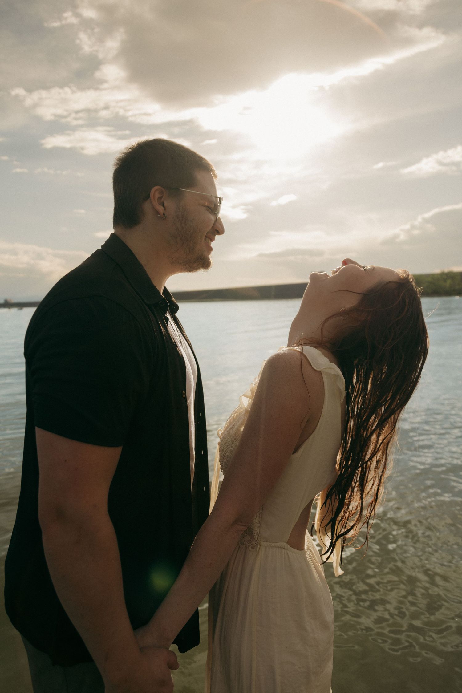 Couple holding hands by a sunlit lake, woman in white dress laughing with hair blowing, man in black shirt smiling.