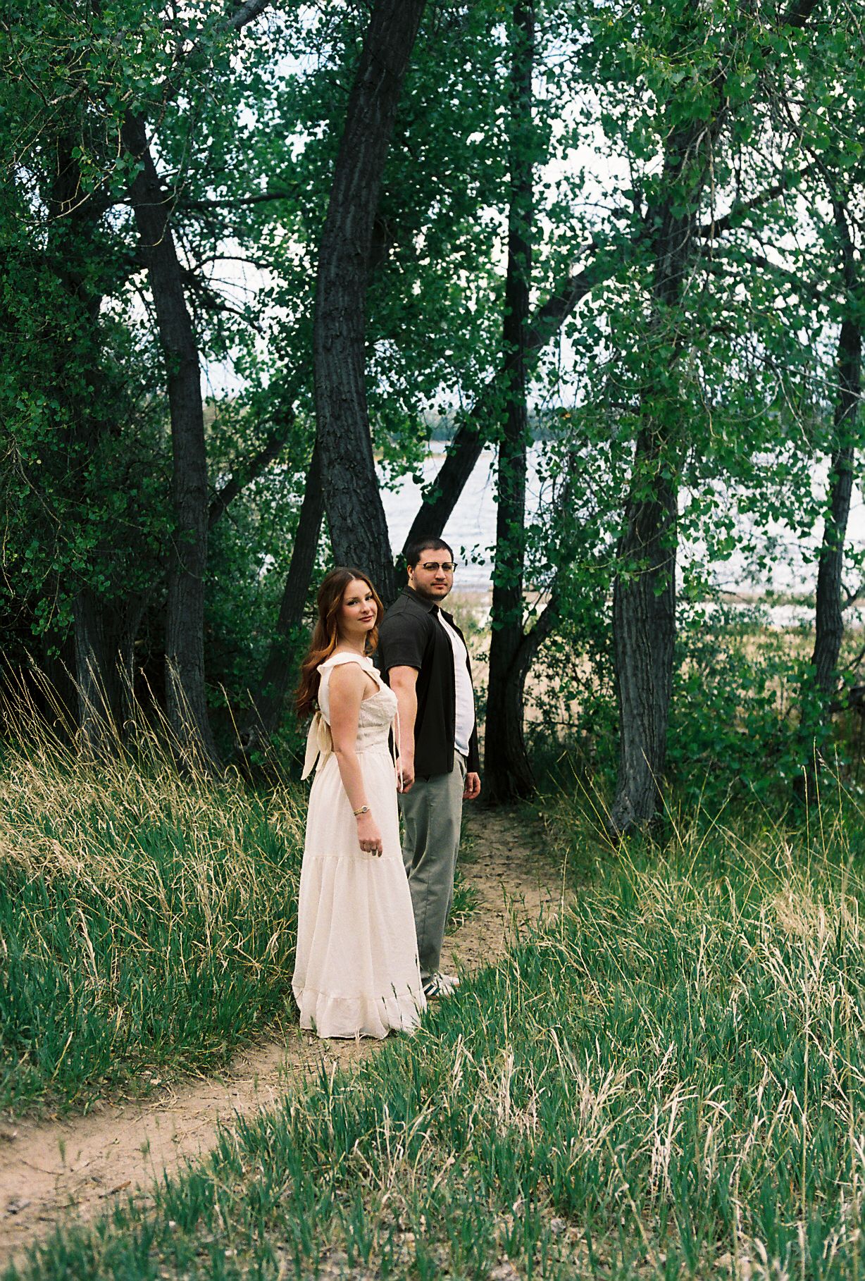A couple stands on a sandy path surrounded by lush green trees, the woman in a white dress and man in casual attire.