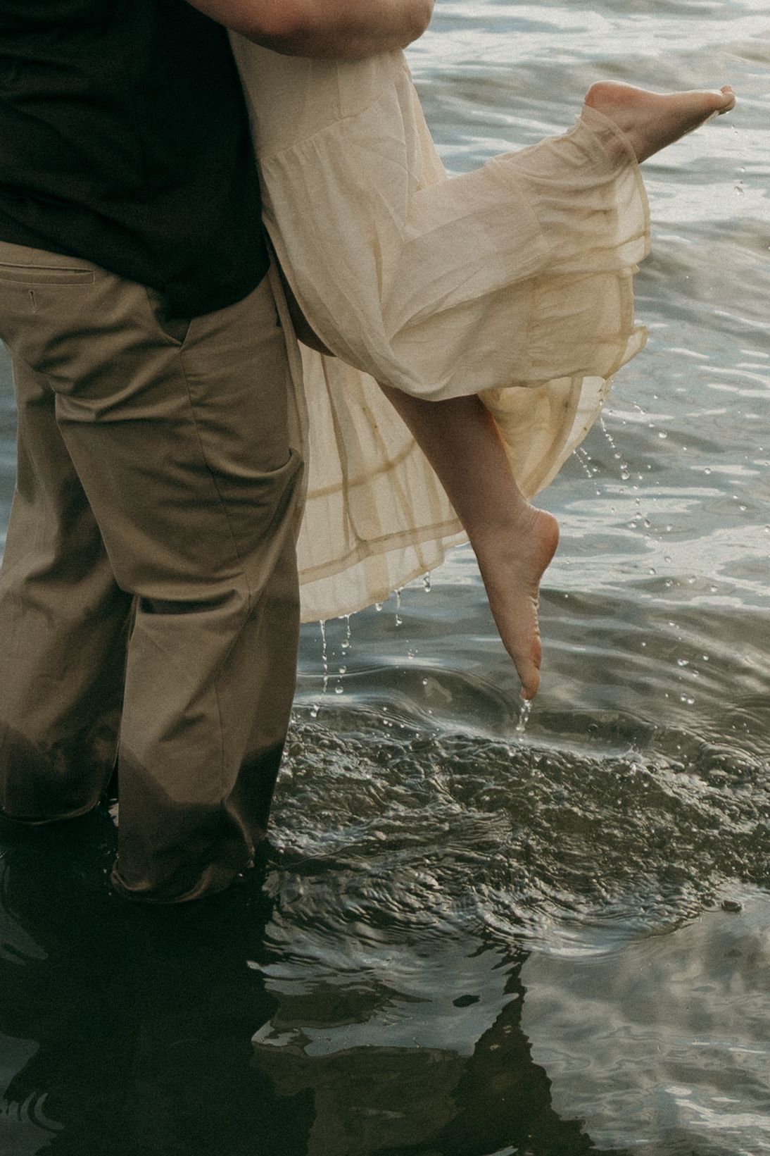 Couple standing in shallow water, woman in flowing cream dress being carried by man in khaki pants near shoreline.