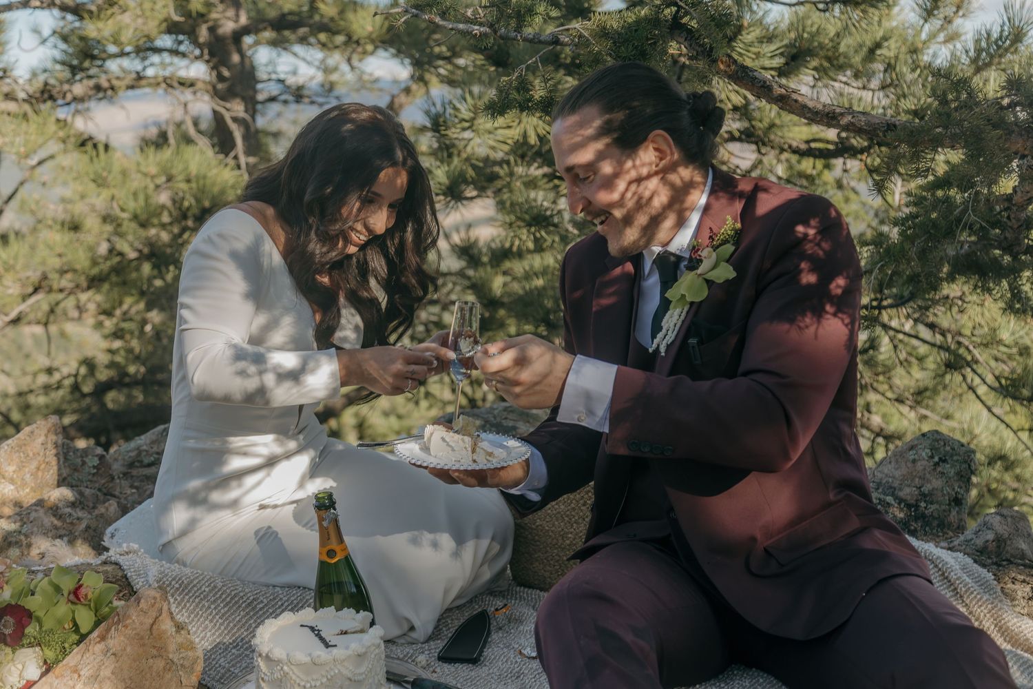 Newlyweds enjoying a champagne picnic celebration among pine trees after their outdoor wedding.