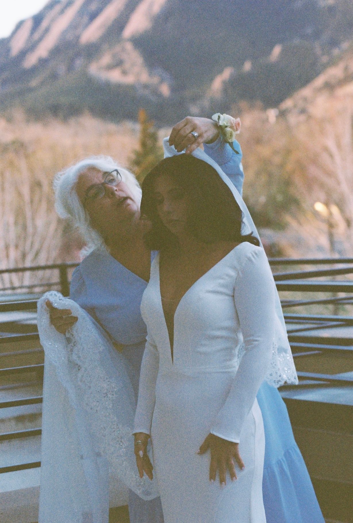 Older woman helps bride with veil on outdoor terrace with Flatirons mountain backdrop.