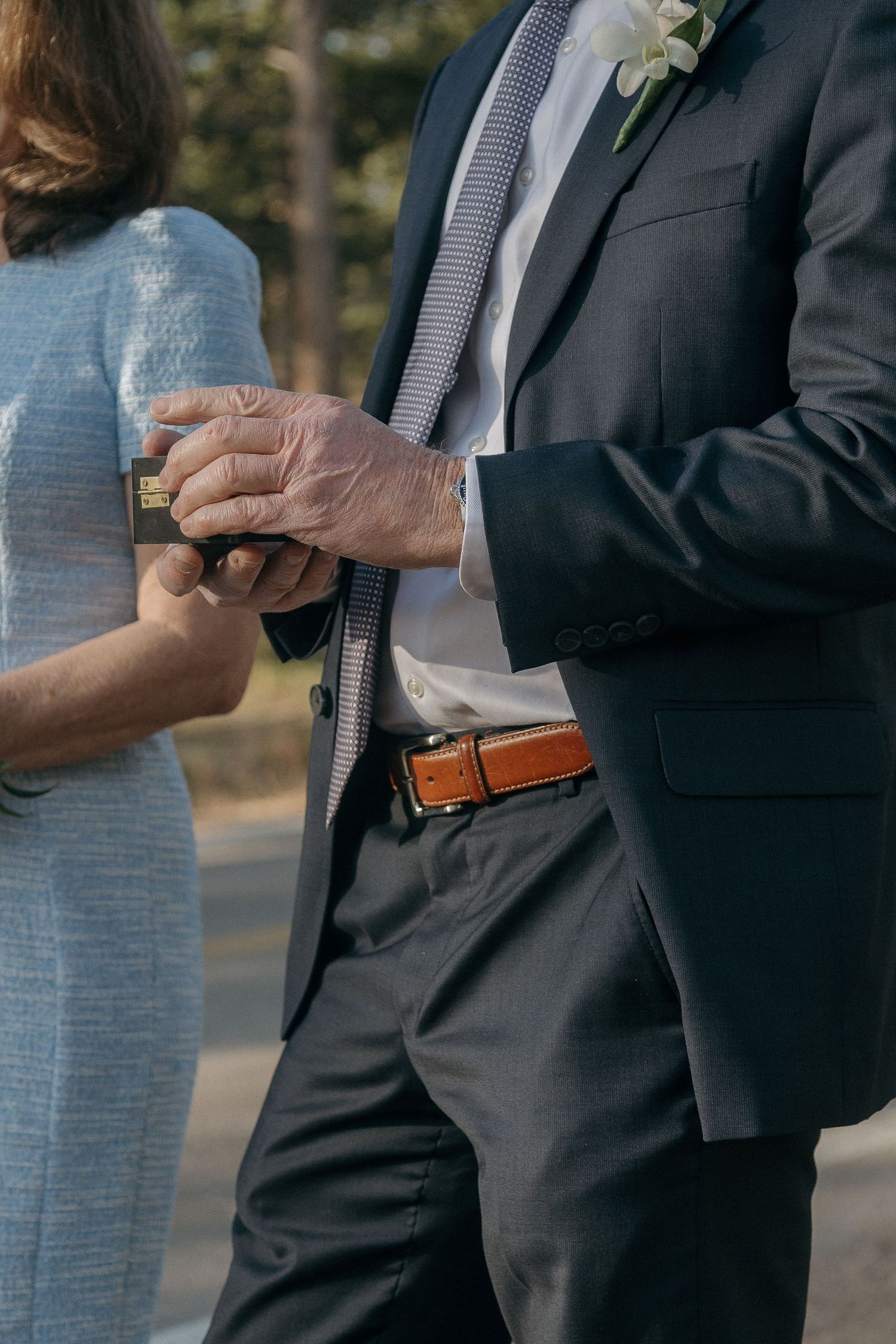 Groom in dark suit with floral boutonniere holds hands with woman in blue dress outdoors.