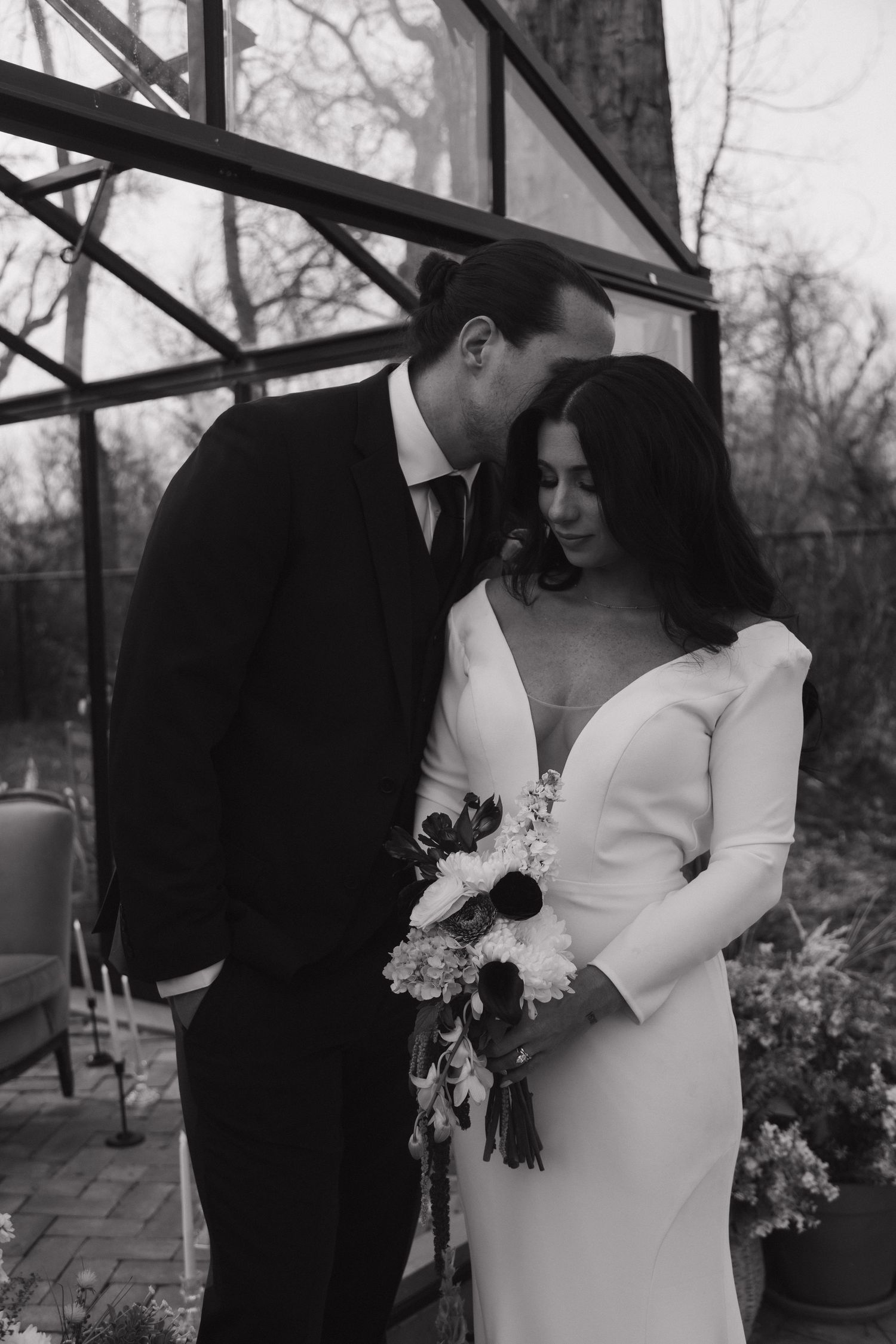 Black and white photo of groom tenderly kissing bride holding a bouquet outside a glass greenhouse.