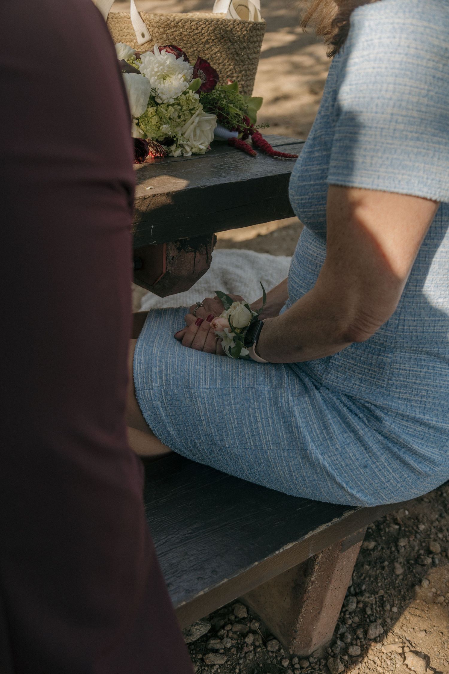 Wedding guest in blue dress holding flowers at outdoor ceremony bench with bouquet of white and green blooms.